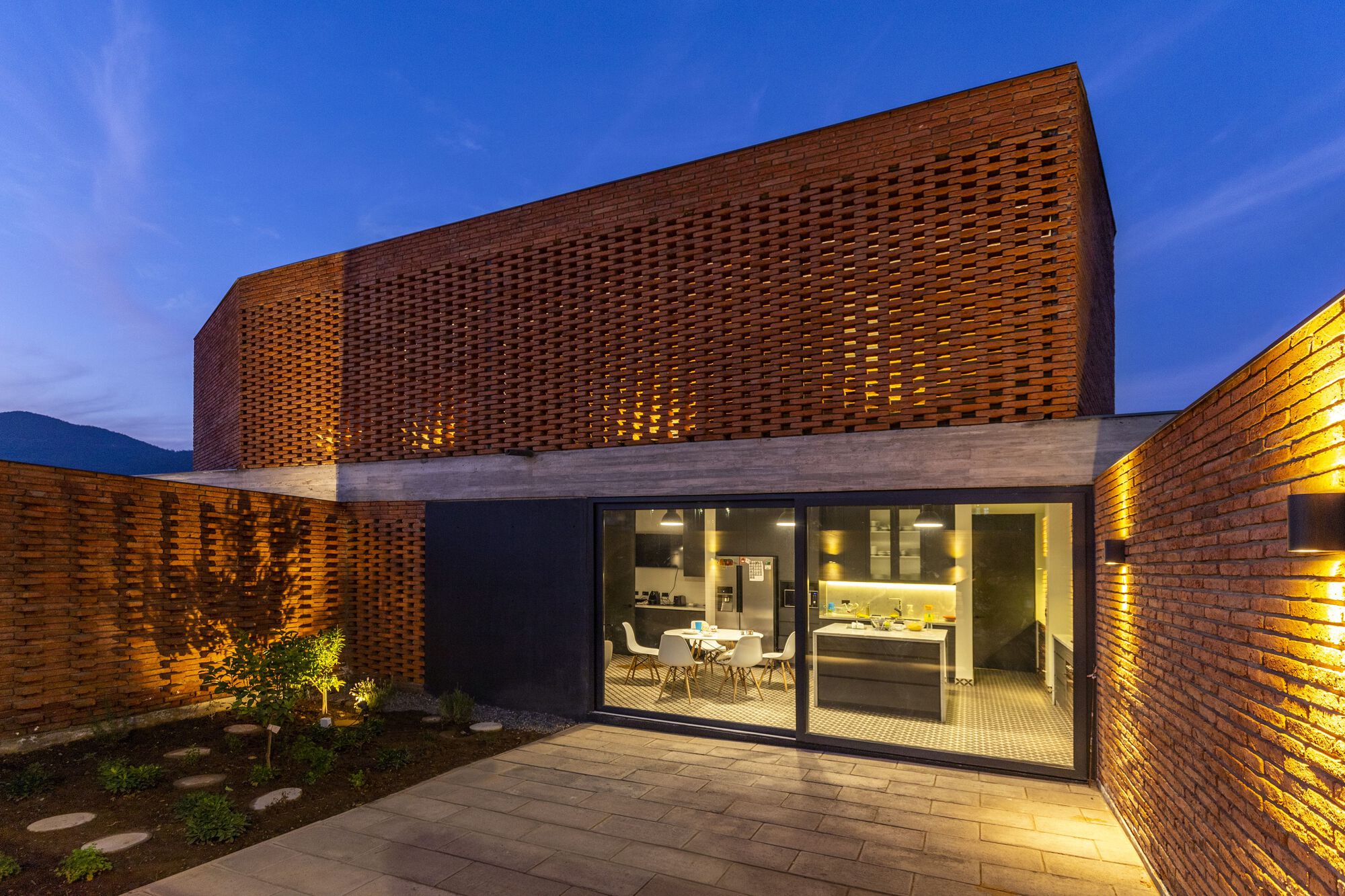 Courtyard view of glazed dining room beneath curved perforated brick volume with planted bed at twilight