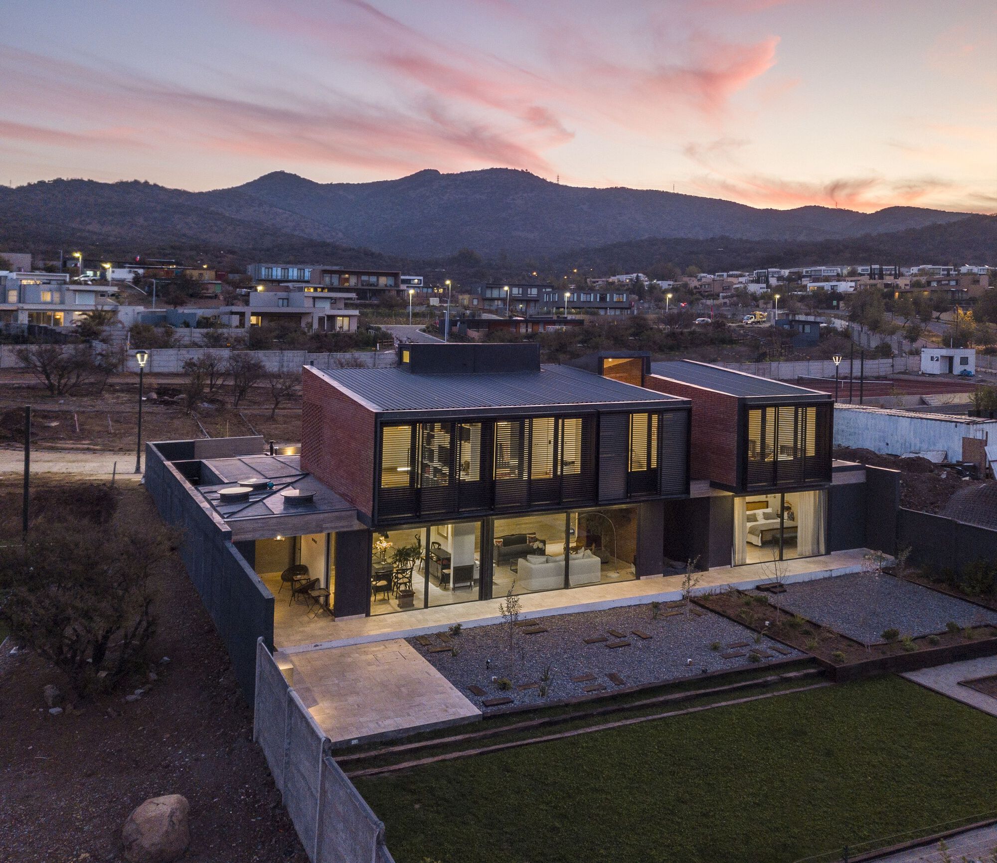 Elevated view of the residence with red brick and glass volumes at dusk with mountains in the background
