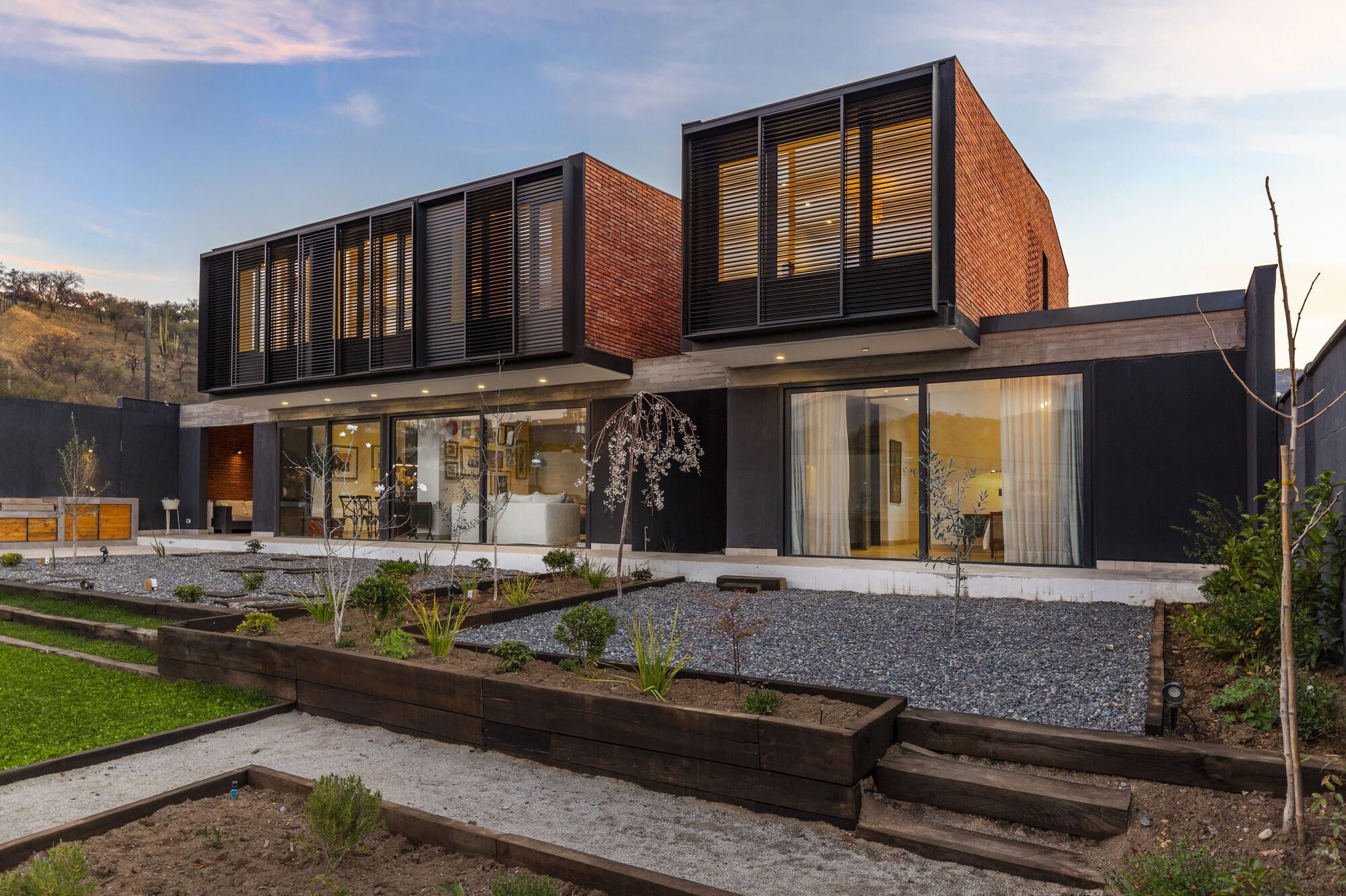 Garden view showing the elevated red brick volumes with horizontal louvers above terraced planted beds and gravel courtyard