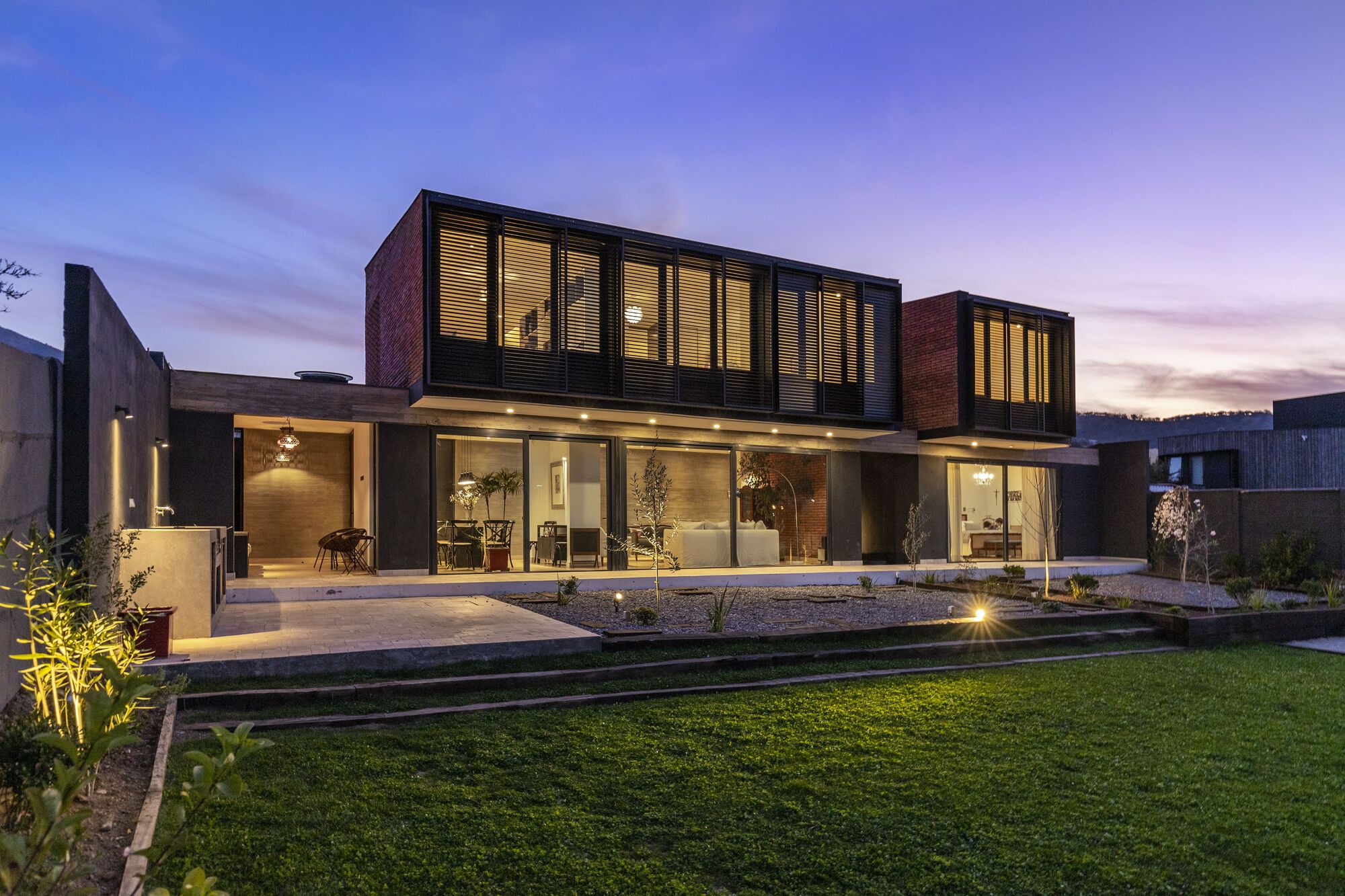 Rear facade at dusk with illuminated glass ground floor and cantilevered brick and metal upper volumes overlooking lawn