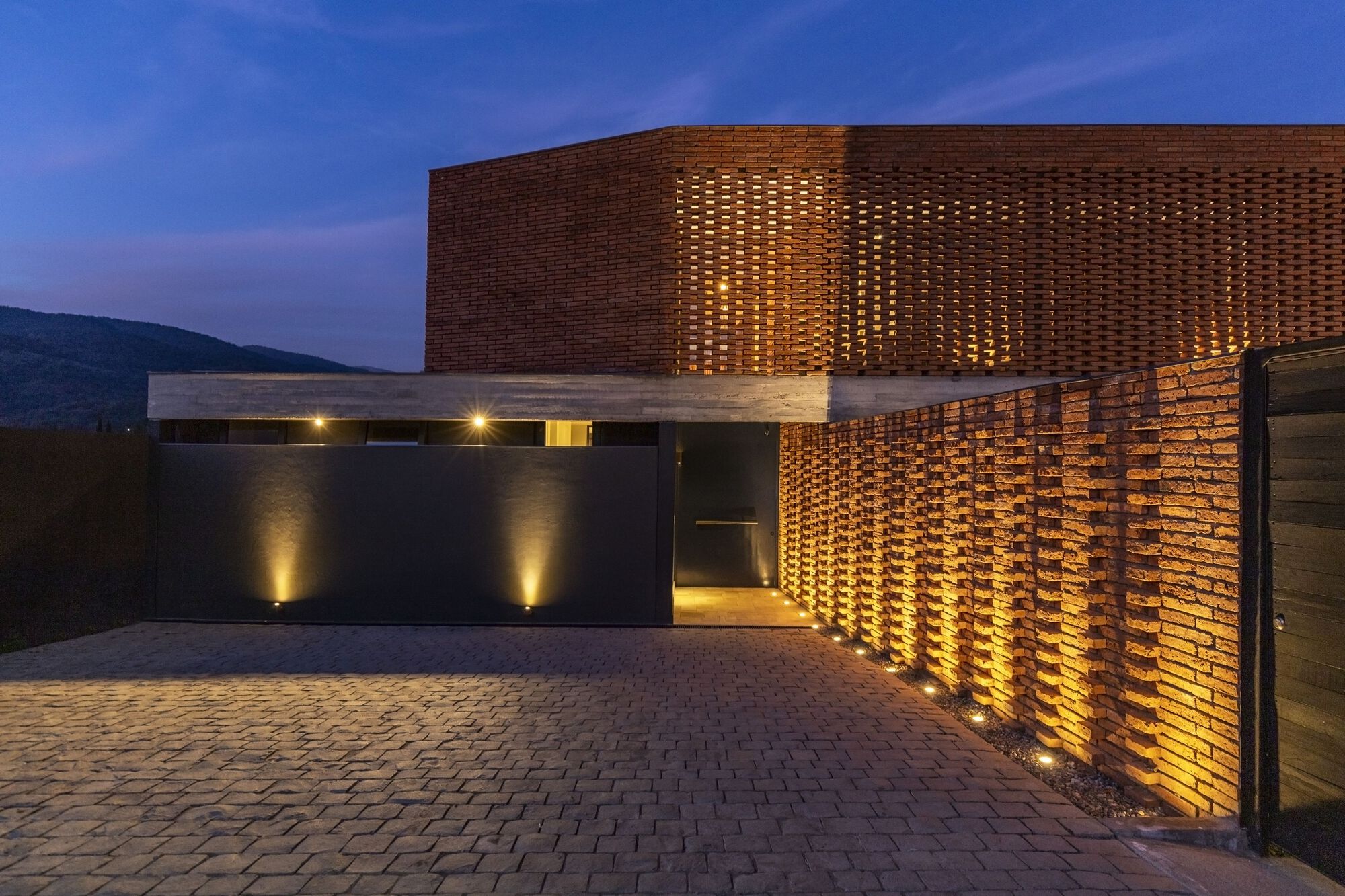 Perforated brick entry wall and canopy lit from below at dusk with paved courtyard