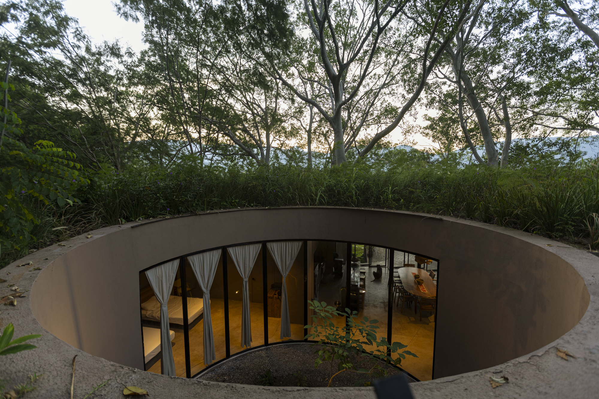 Circular courtyard with glazed doors opening to the interior beneath a grass-covered roof edge at twilight