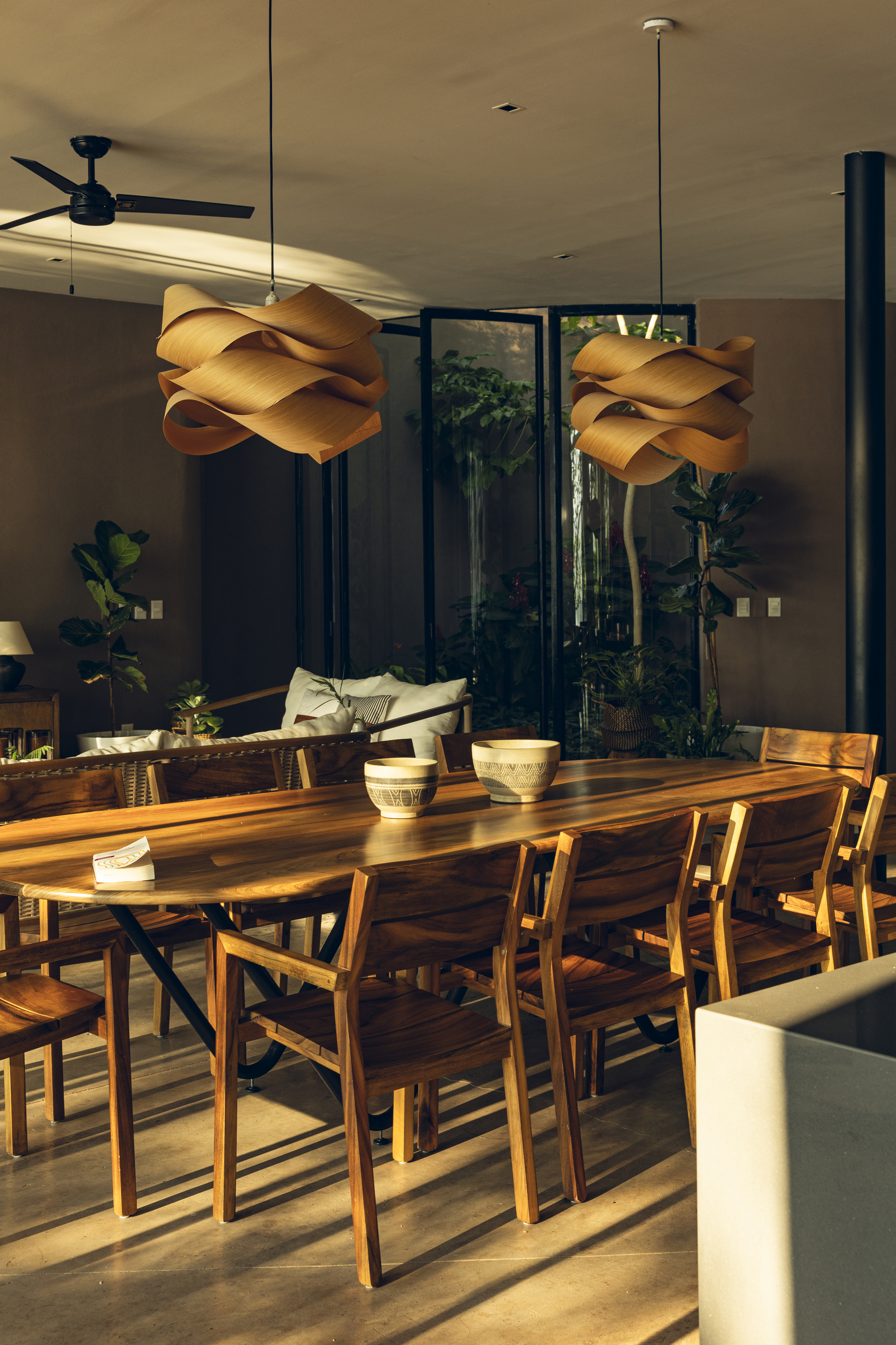 Timber dining table beneath sculptural pendant lights with afternoon sun casting shadows across the concrete floor