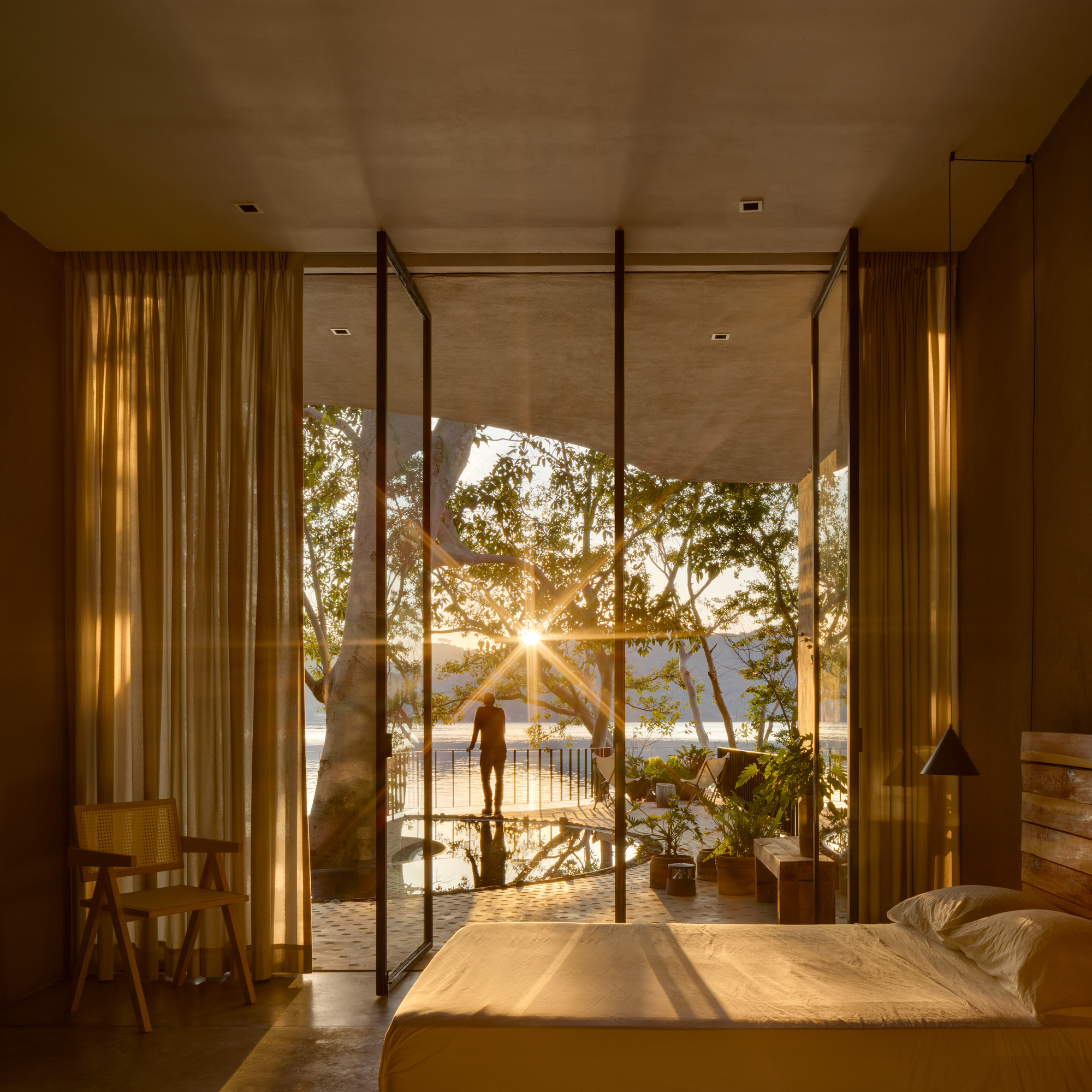 Bedroom with steel-framed glass doors opening to a terrace with a large tree at sunset