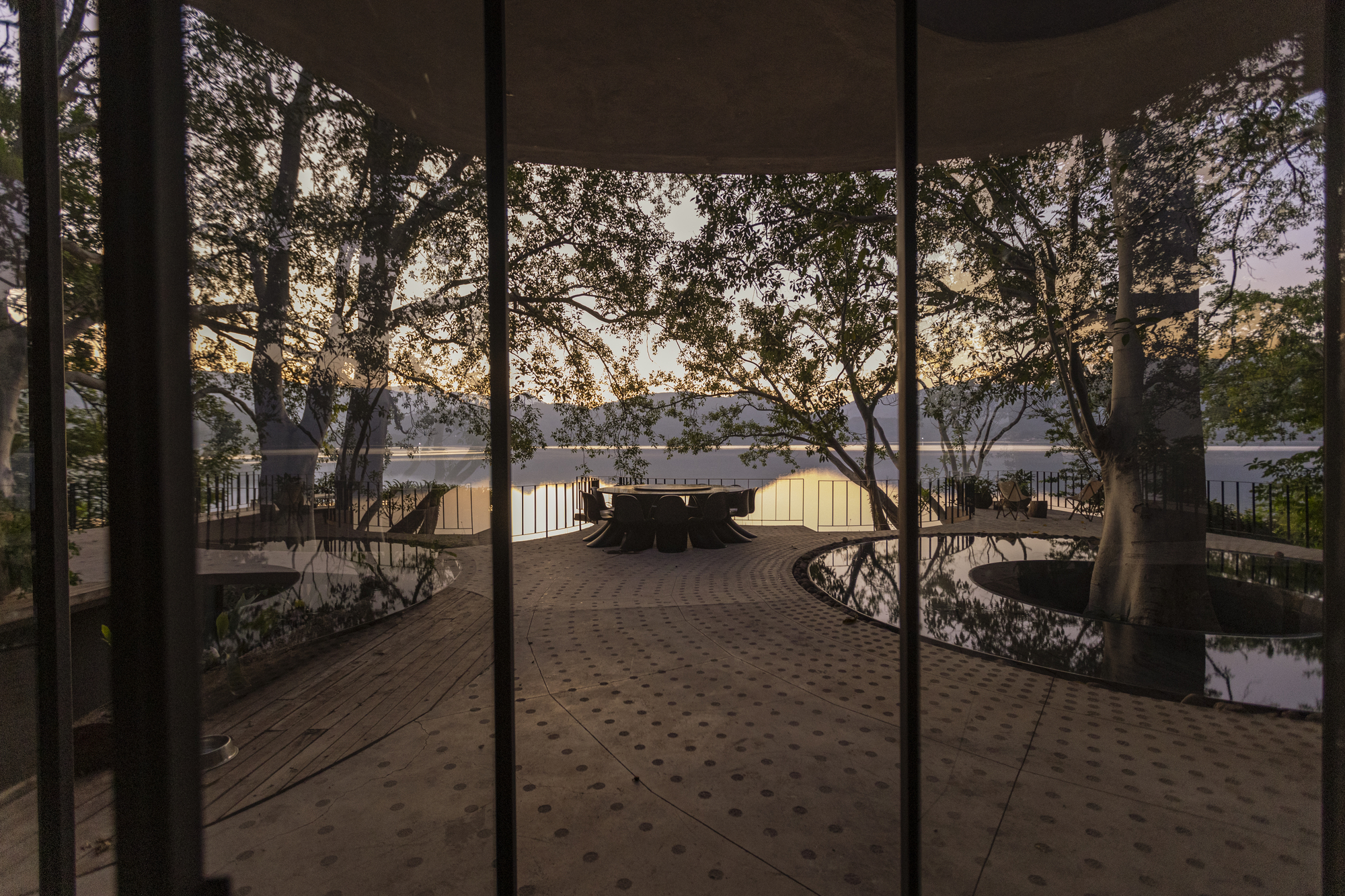 View through glass doors to an elevated timber deck with preserved trees and a distant lake at dusk