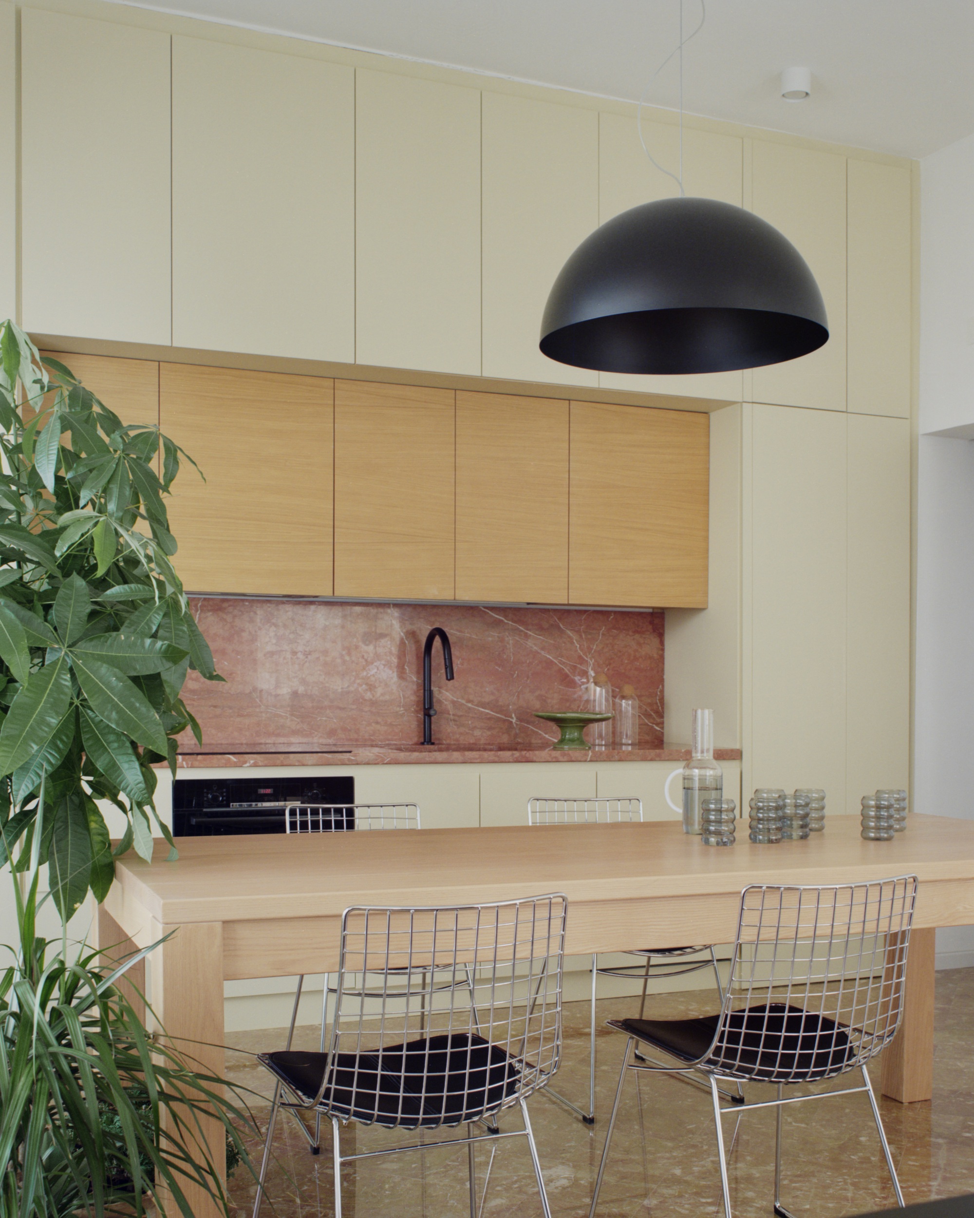 Kitchen with oak and cream cabinetry, pink marble backsplash and wire mesh dining chairs beneath a dome pendant