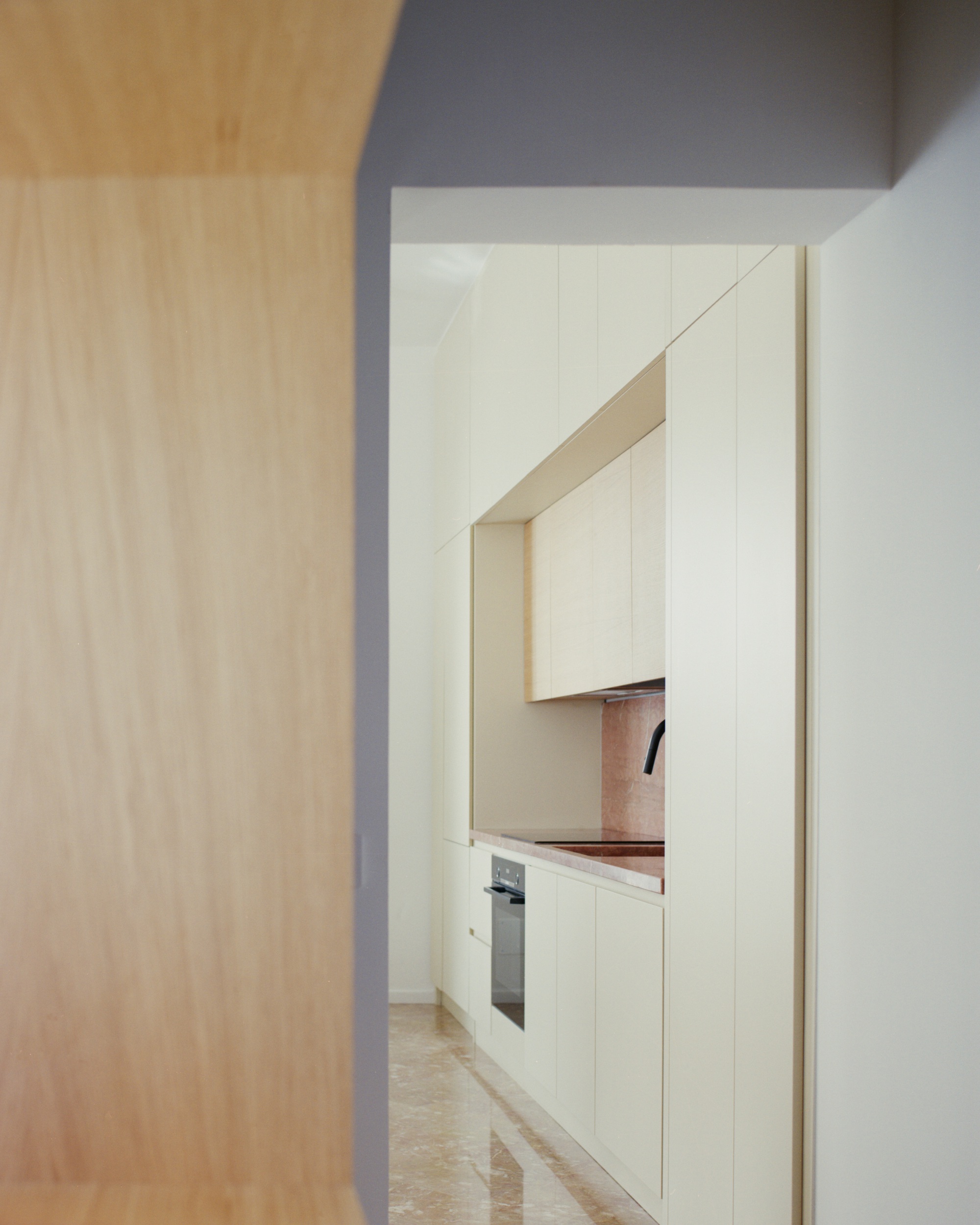 Doorway framed by pale wood paneling revealing kitchen with pink marble countertop and cream upper cabinets