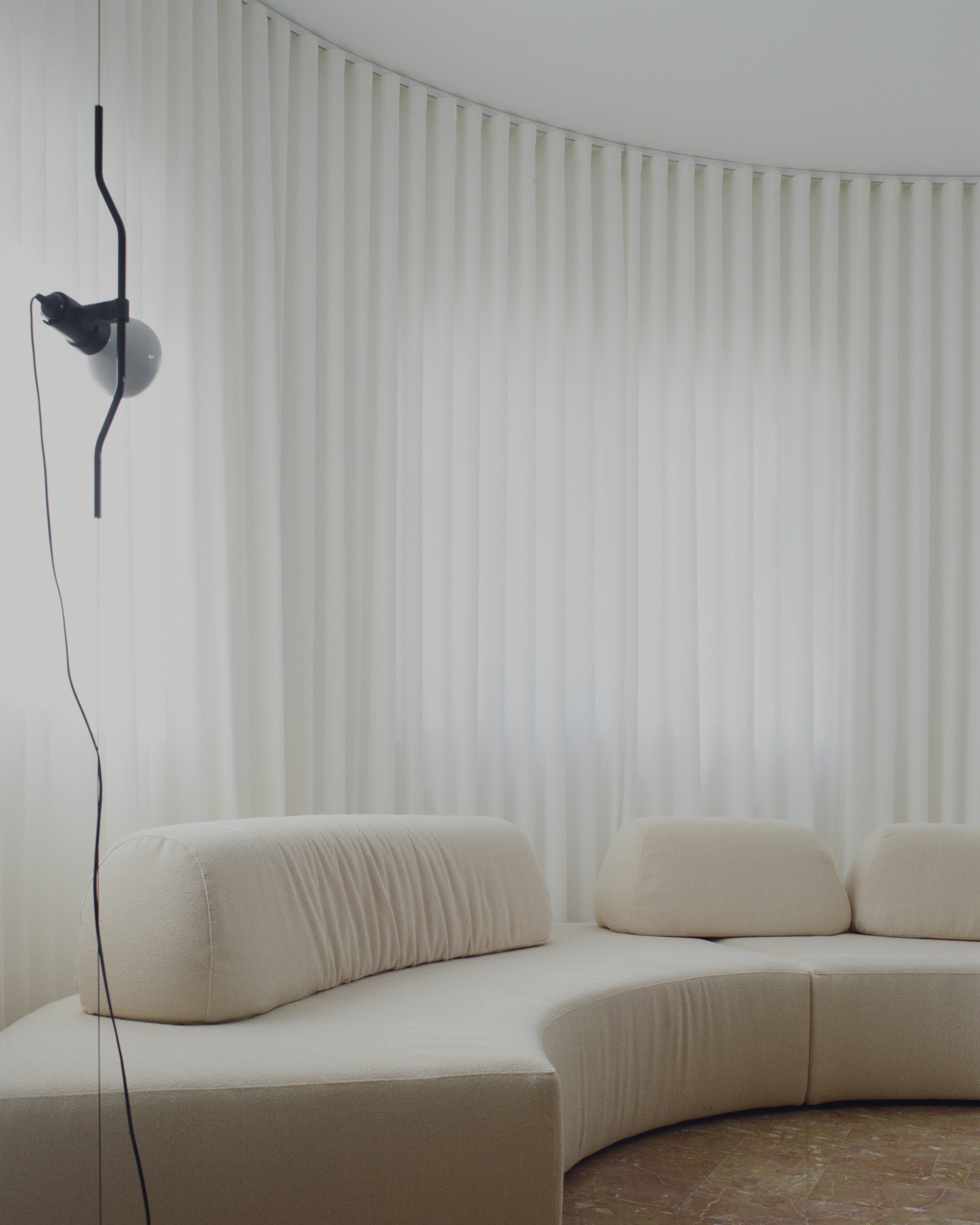 Curved seating upholstered in pale fabric against floor-to-ceiling curtains with a wall-mounted black reading lamp