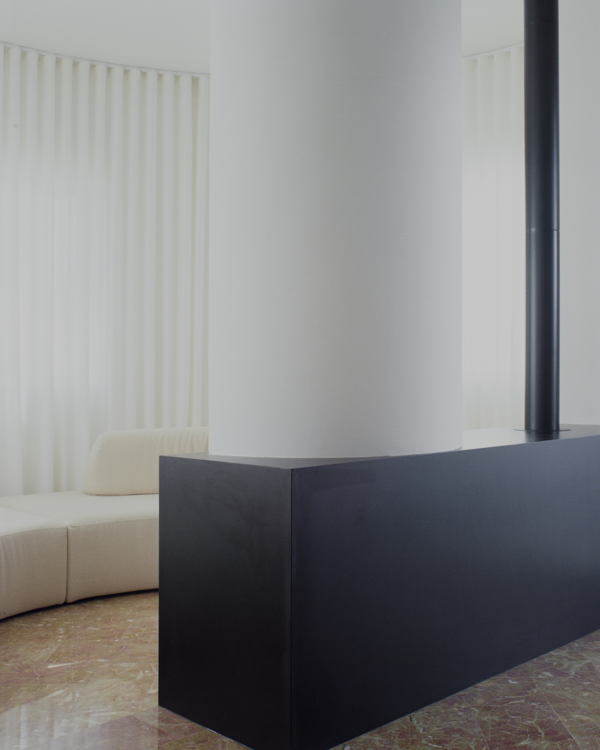 Black steel fireplace hearth next to curved sofa with white curtain and stone floor