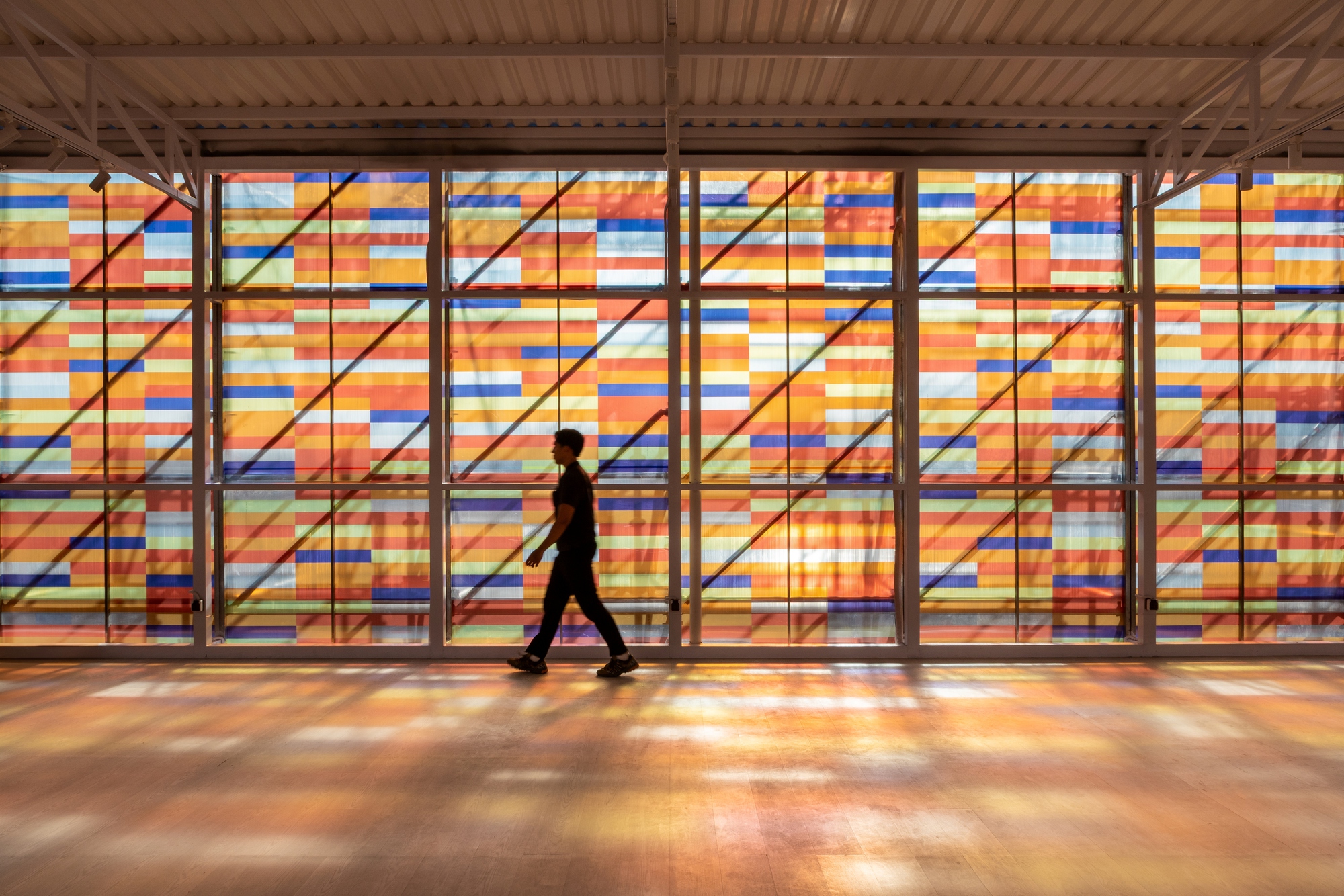 Interior view of a person walking past the backlit striped panels filtering daylight through timber framing