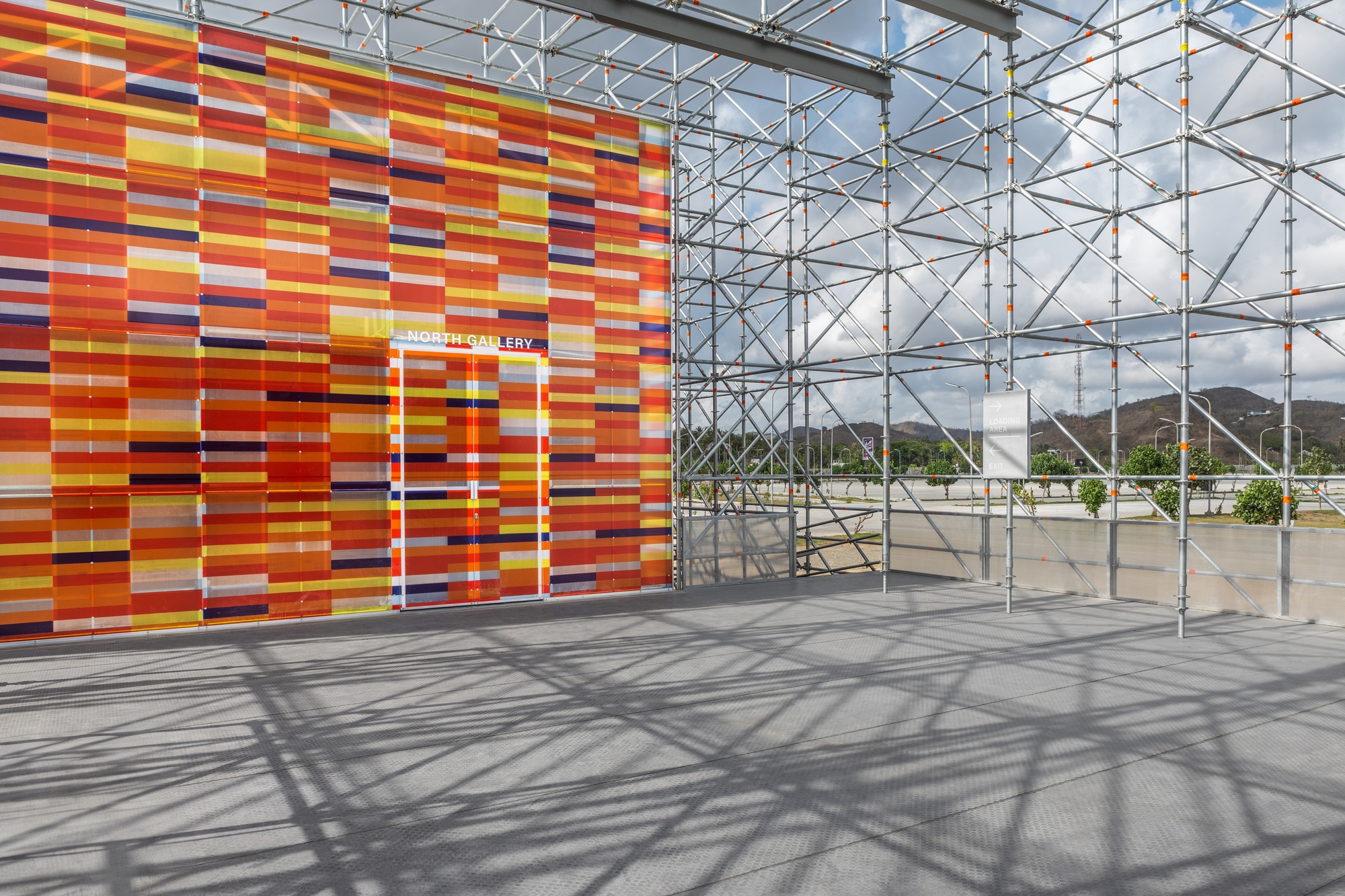 Entry doorway cut through the multicolored horizontal panel wall with scaffolding casting shadows on concrete