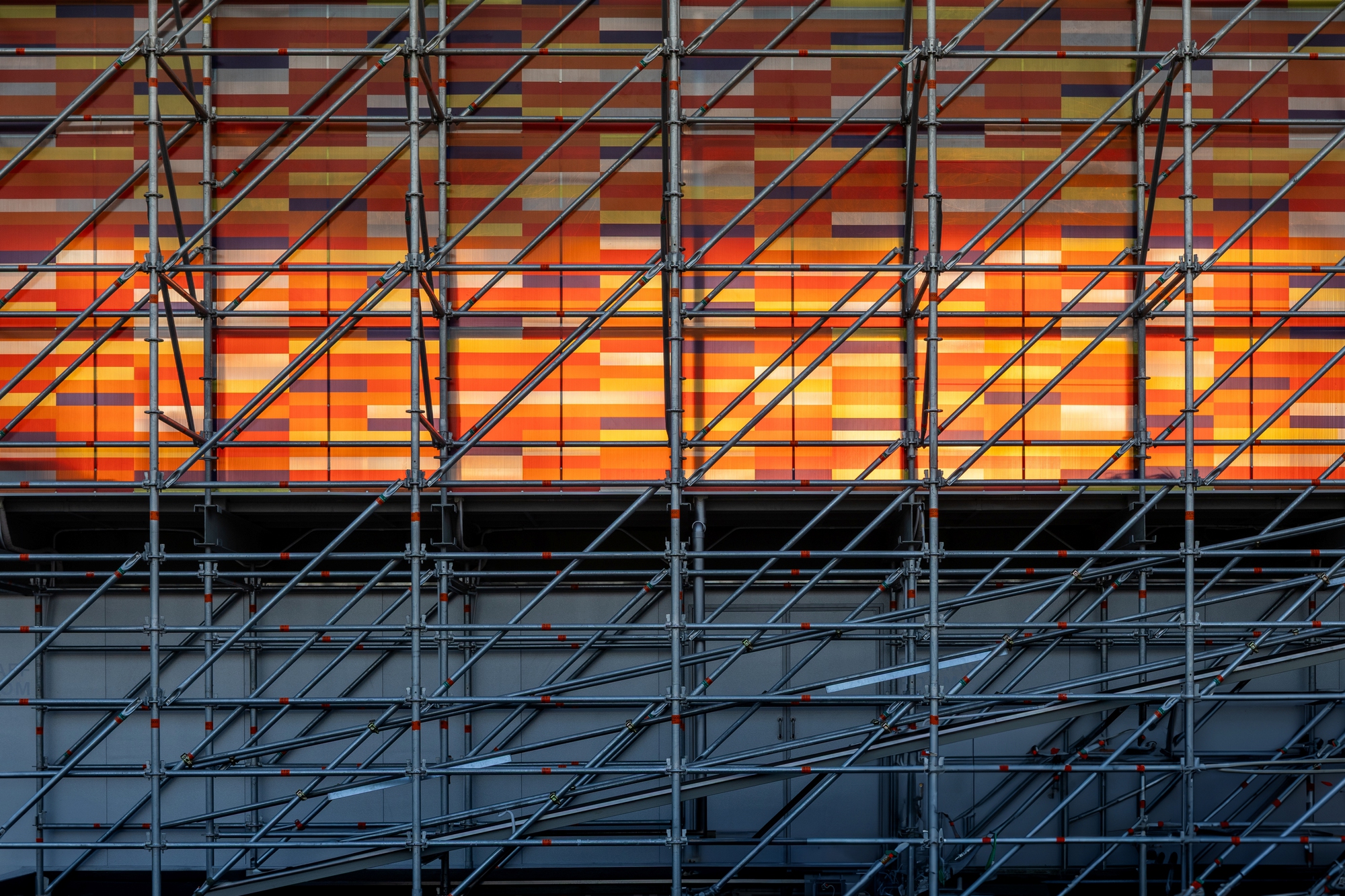 Horizontal bands of orange and warm-toned panels backlit behind the diagonal steel cross-bracing