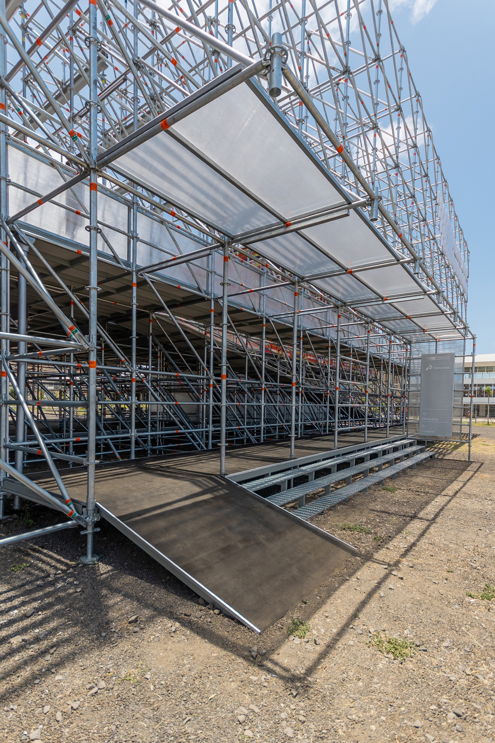 Metal scaffold structure with tensile fabric canopy and accessible ramp under clear blue sky