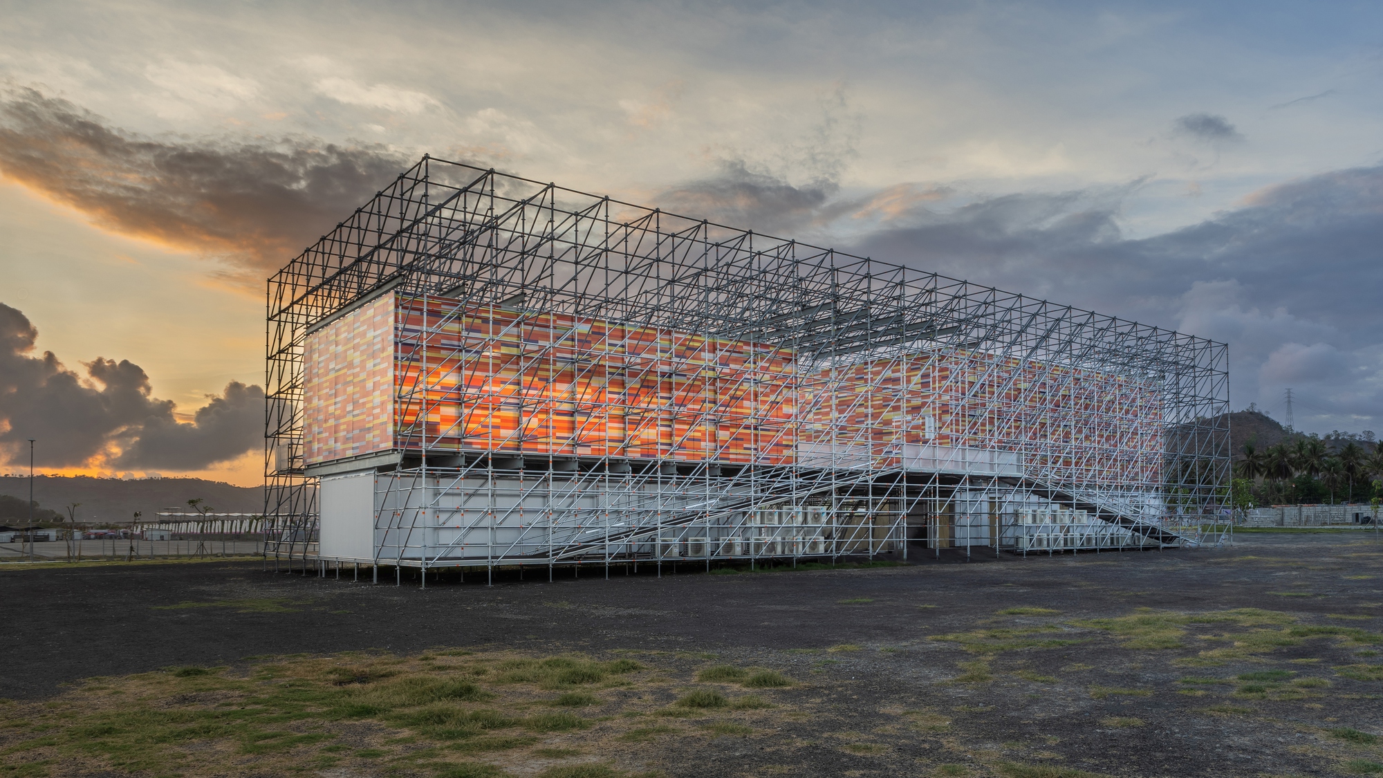 Steel frame pavilion with translucent orange panels glowing at dusk beneath dramatic storm clouds