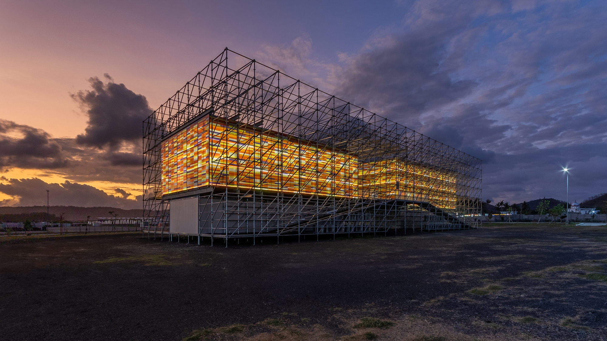 Illuminated scaffold-clad structure with glowing amber facade at twilight against purple and orange sky