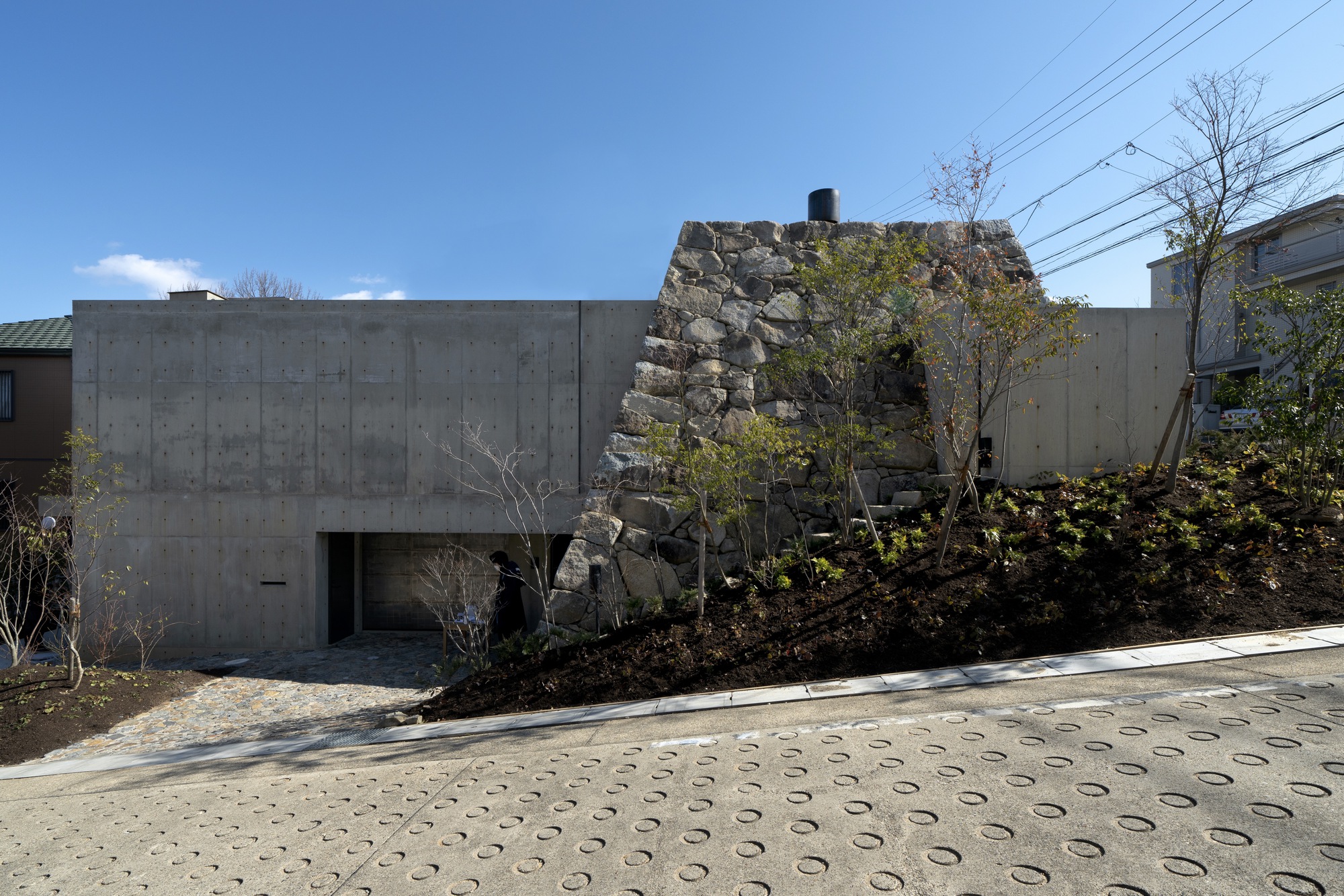 Concrete facade with recessed entrance beside a reconstructed stone wall and paved plaza under blue sky