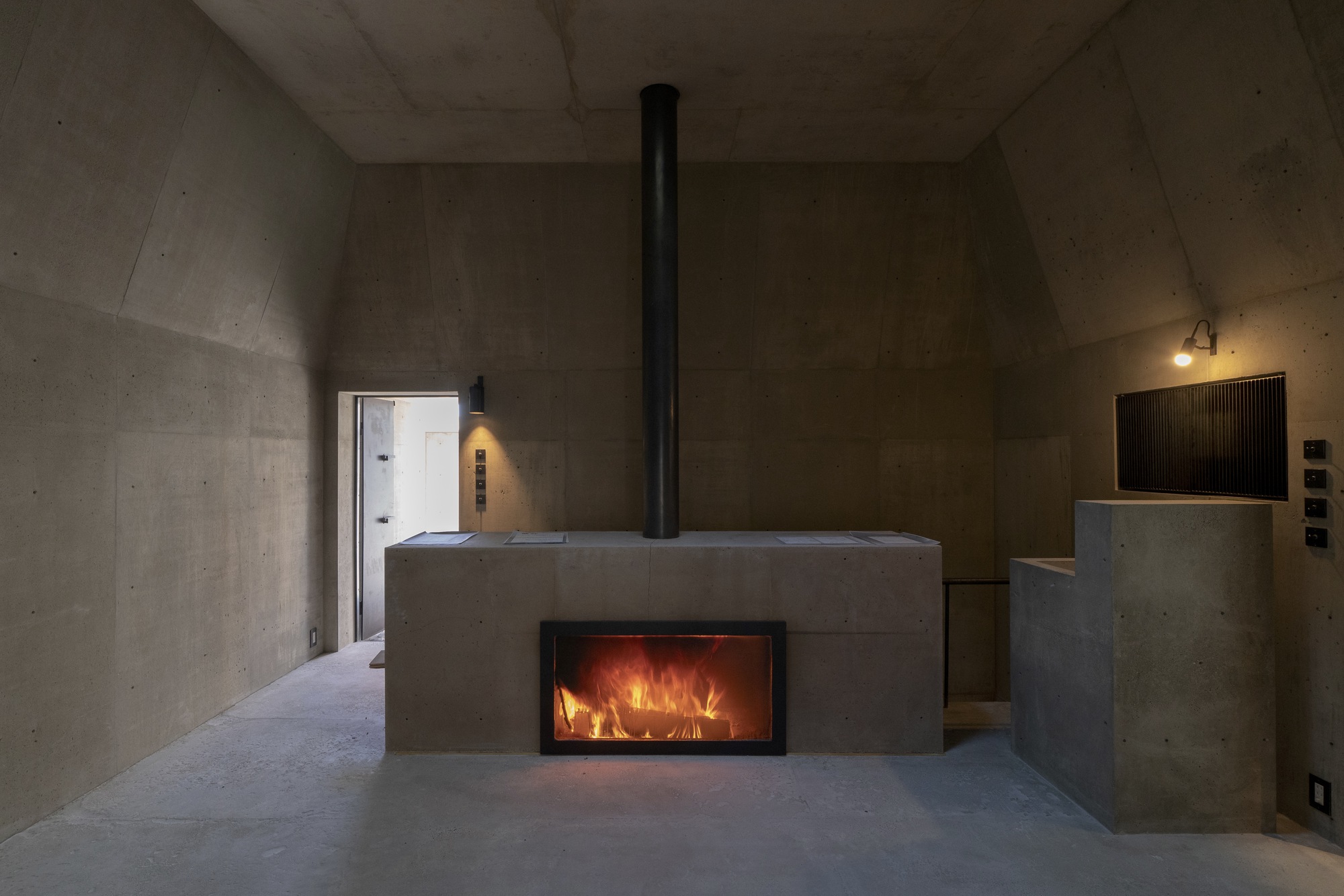 Concrete fireplace block with black flue pipe and glowing fire inside a vaulted room at dusk