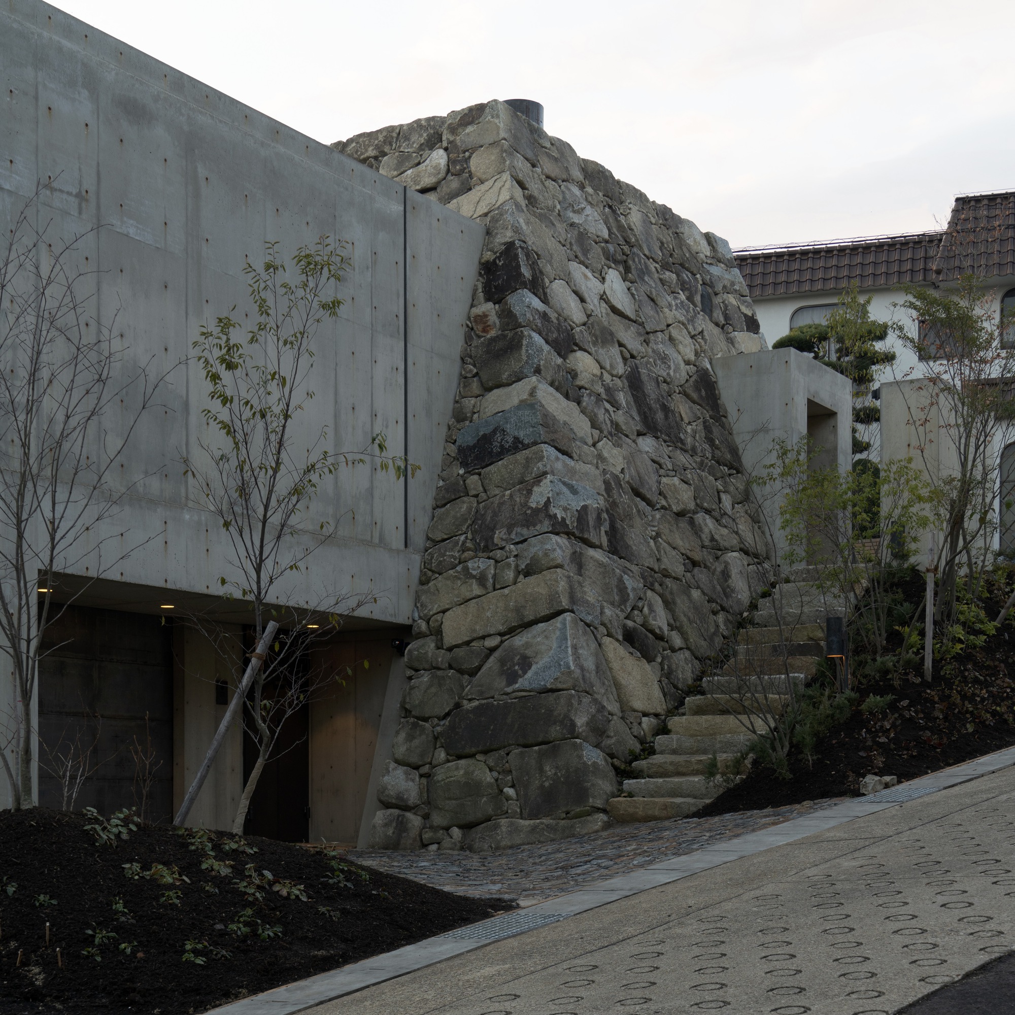 Exposed stone outcrop integrated into a concrete facade with planted beds and young trees below
