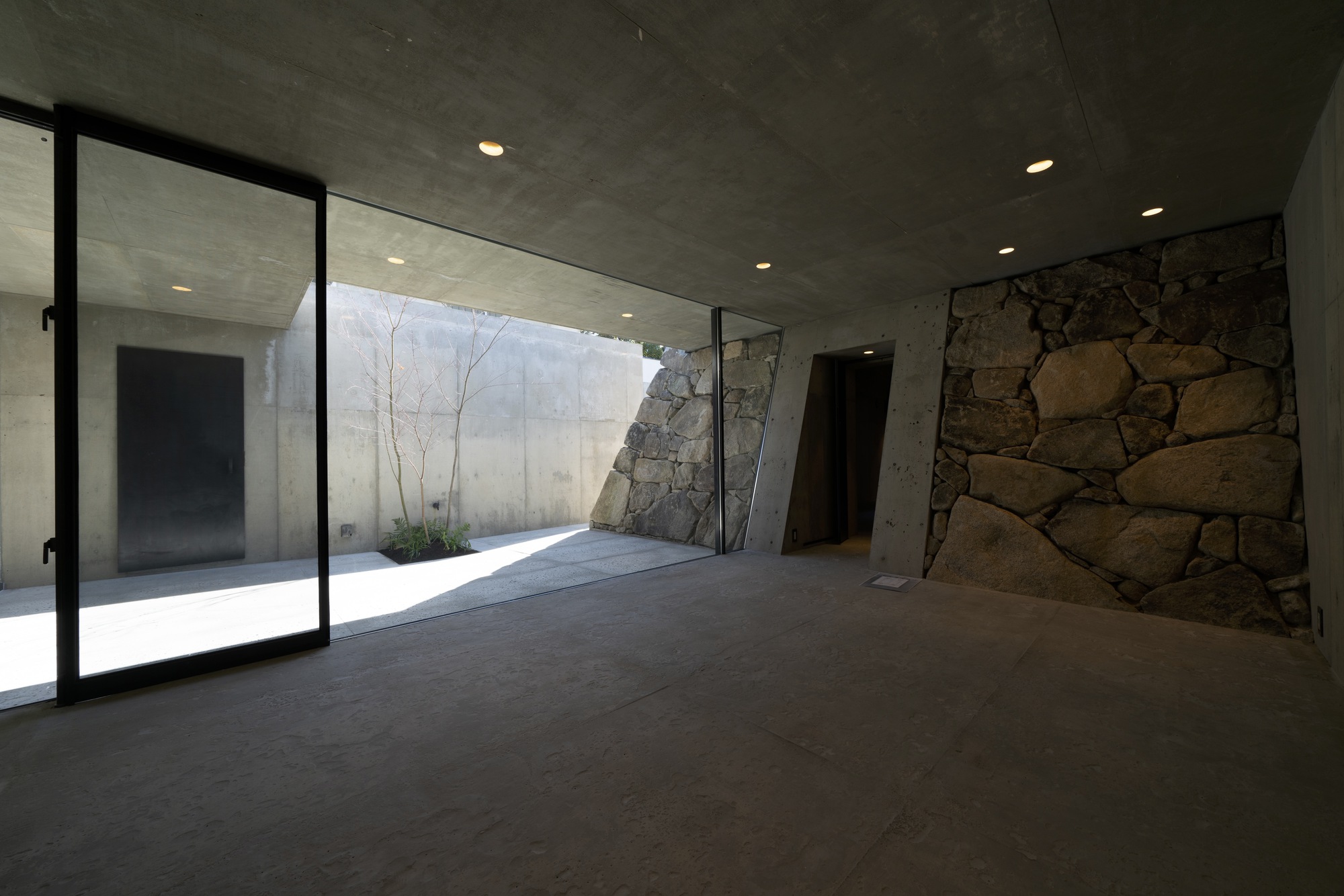 Interior space with concrete ceiling and floor-to-ceiling glass opening onto a stone-walled courtyard