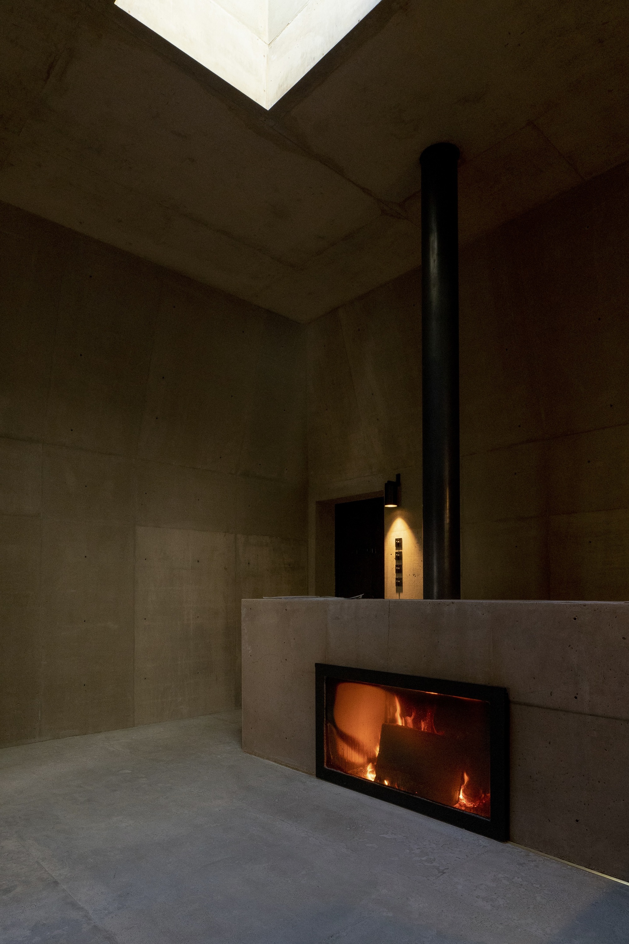 Glass-fronted fireplace set into a concrete volume beneath a black metal flue and skylight