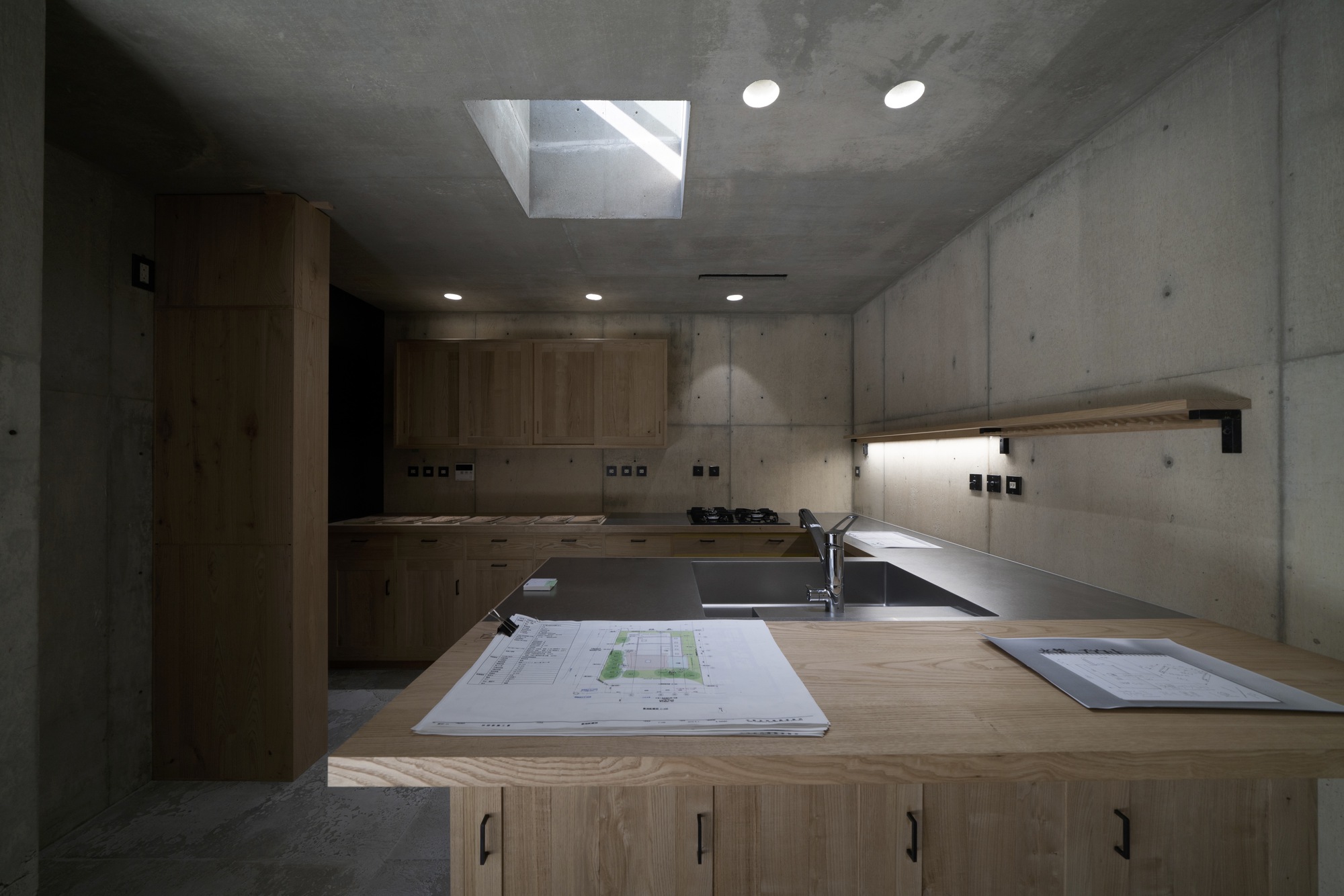 Kitchen with timber cabinetry and board-formed concrete walls illuminated by a central skylight