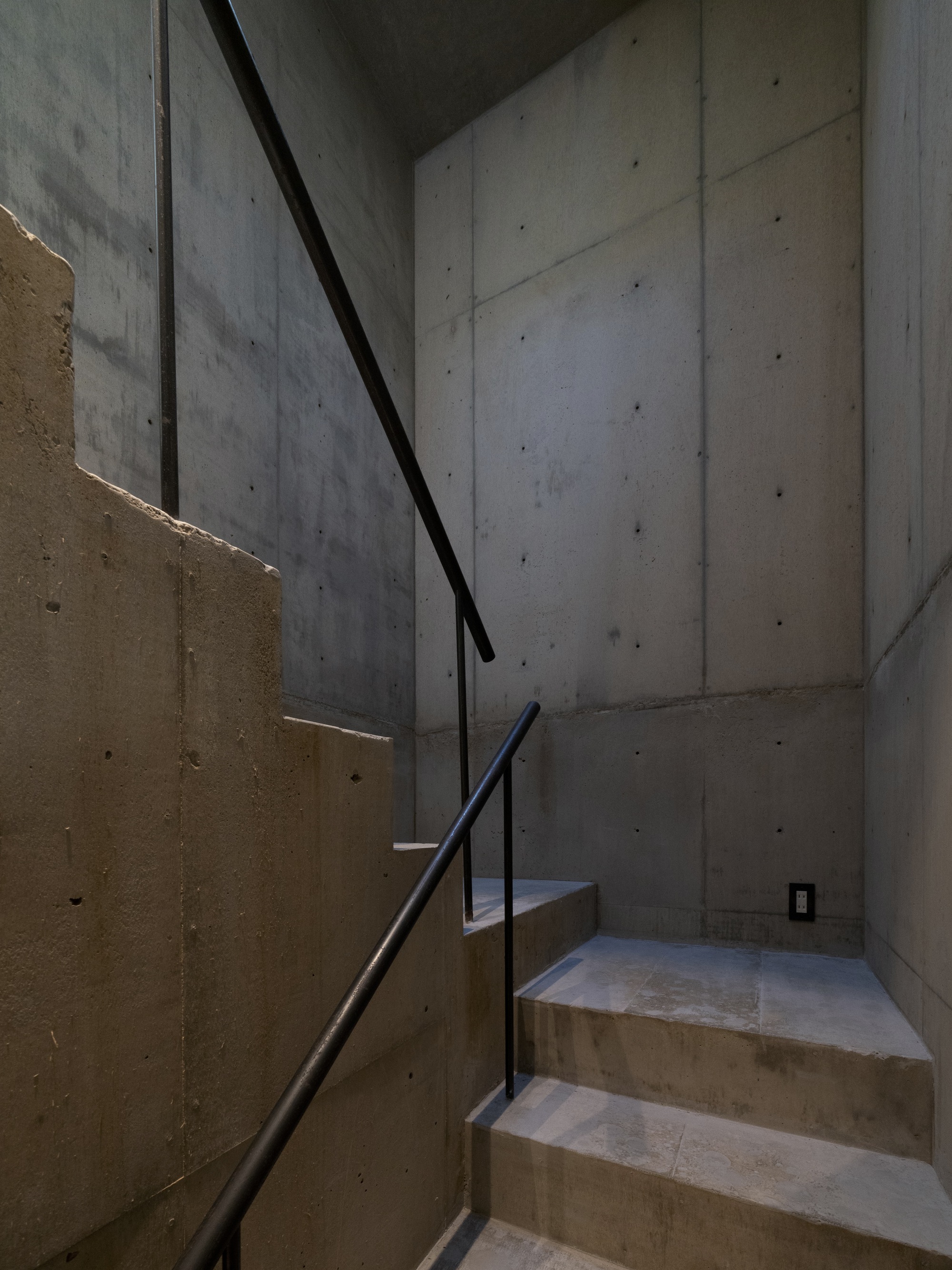 Board-formed concrete staircase with black steel handrail ascending between cast concrete walls