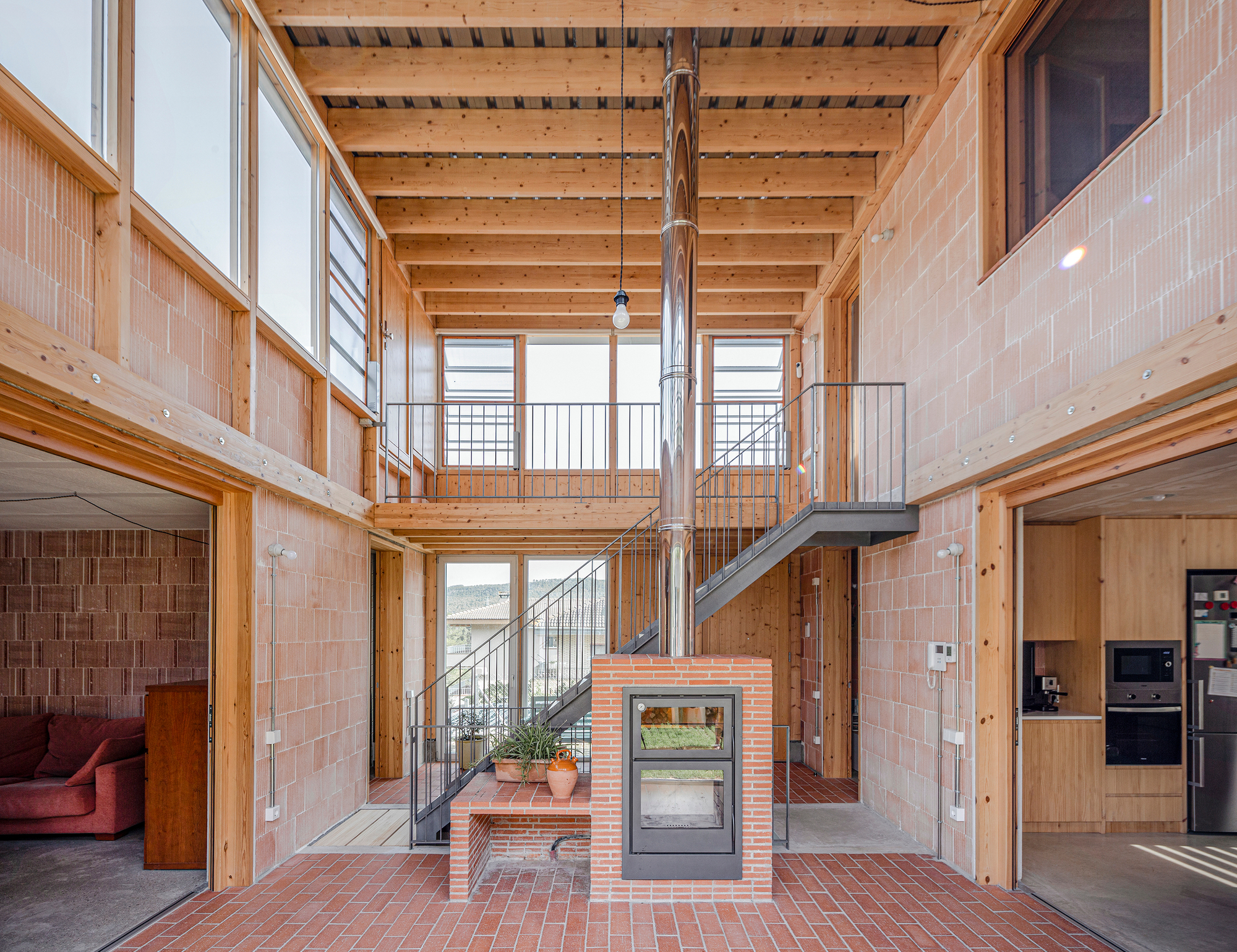 Double-height interior with exposed timber beams and a freestanding brick fireplace beneath a steel flue pipe
