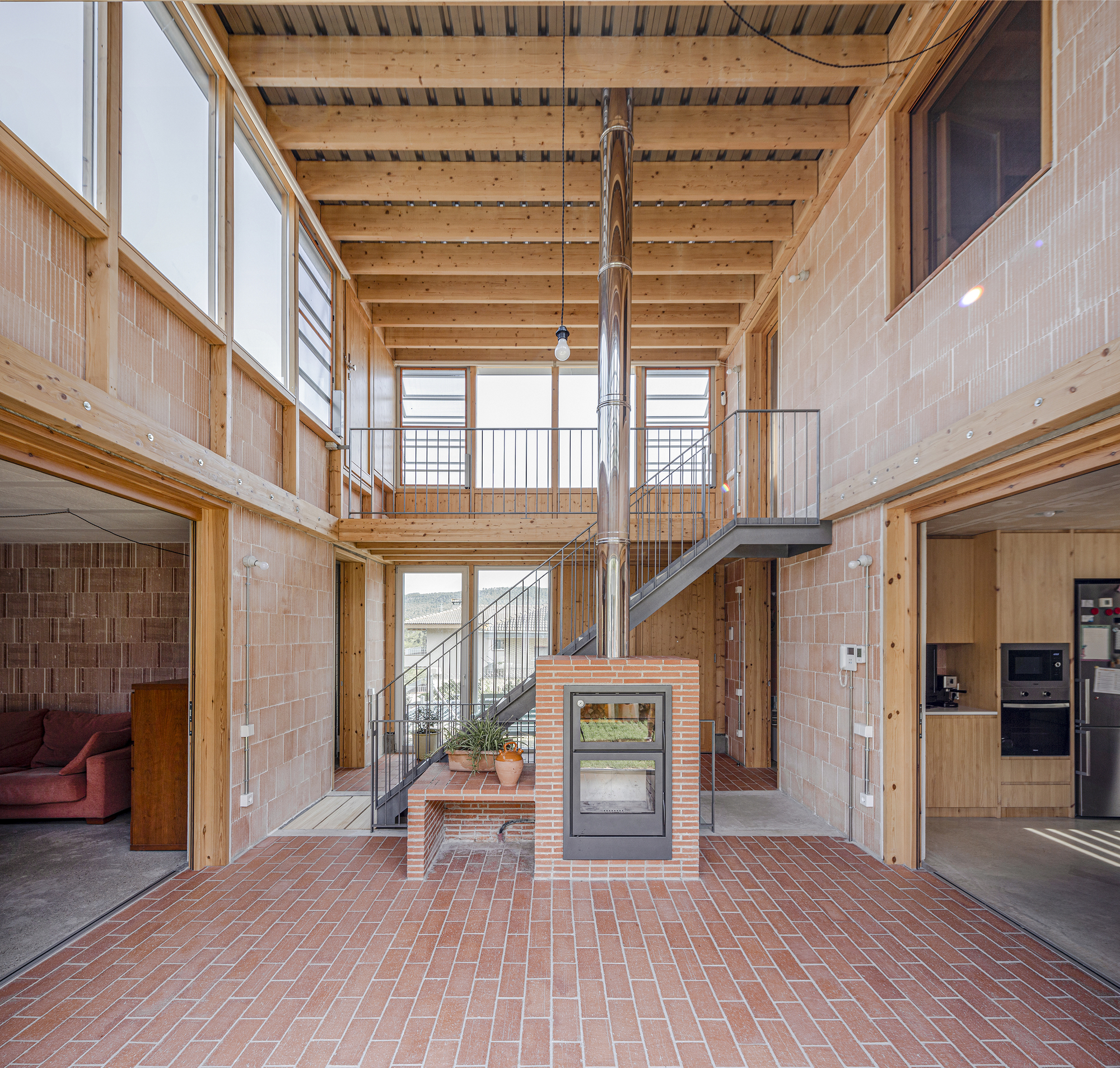 Central atrium with timber ceiling structure, steel staircase, and freestanding brick fireplace on herringbone pavers