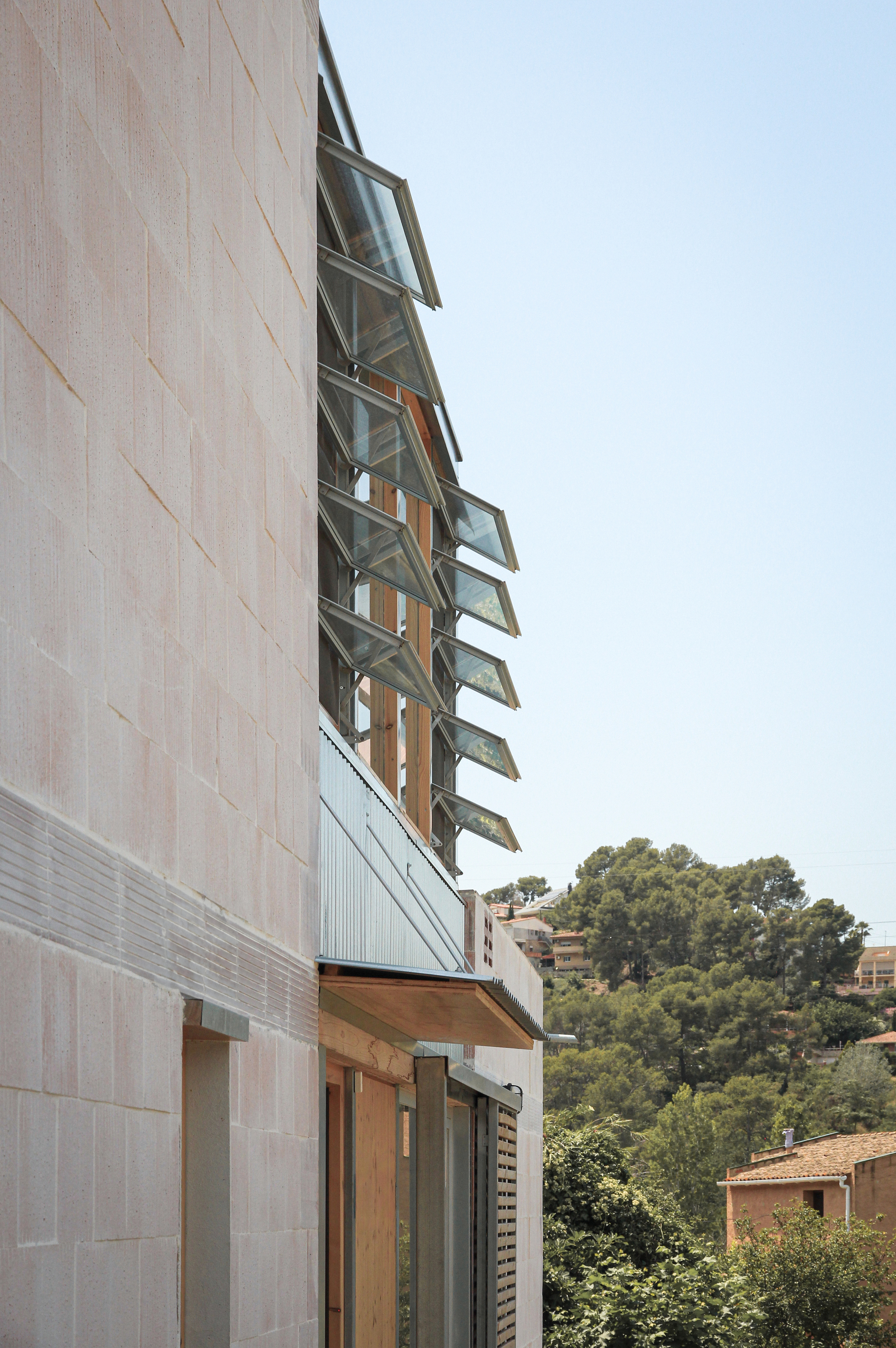 Exterior facade with vertically stacked operable glass windows set between terracotta tile cladding and hillside vegetation