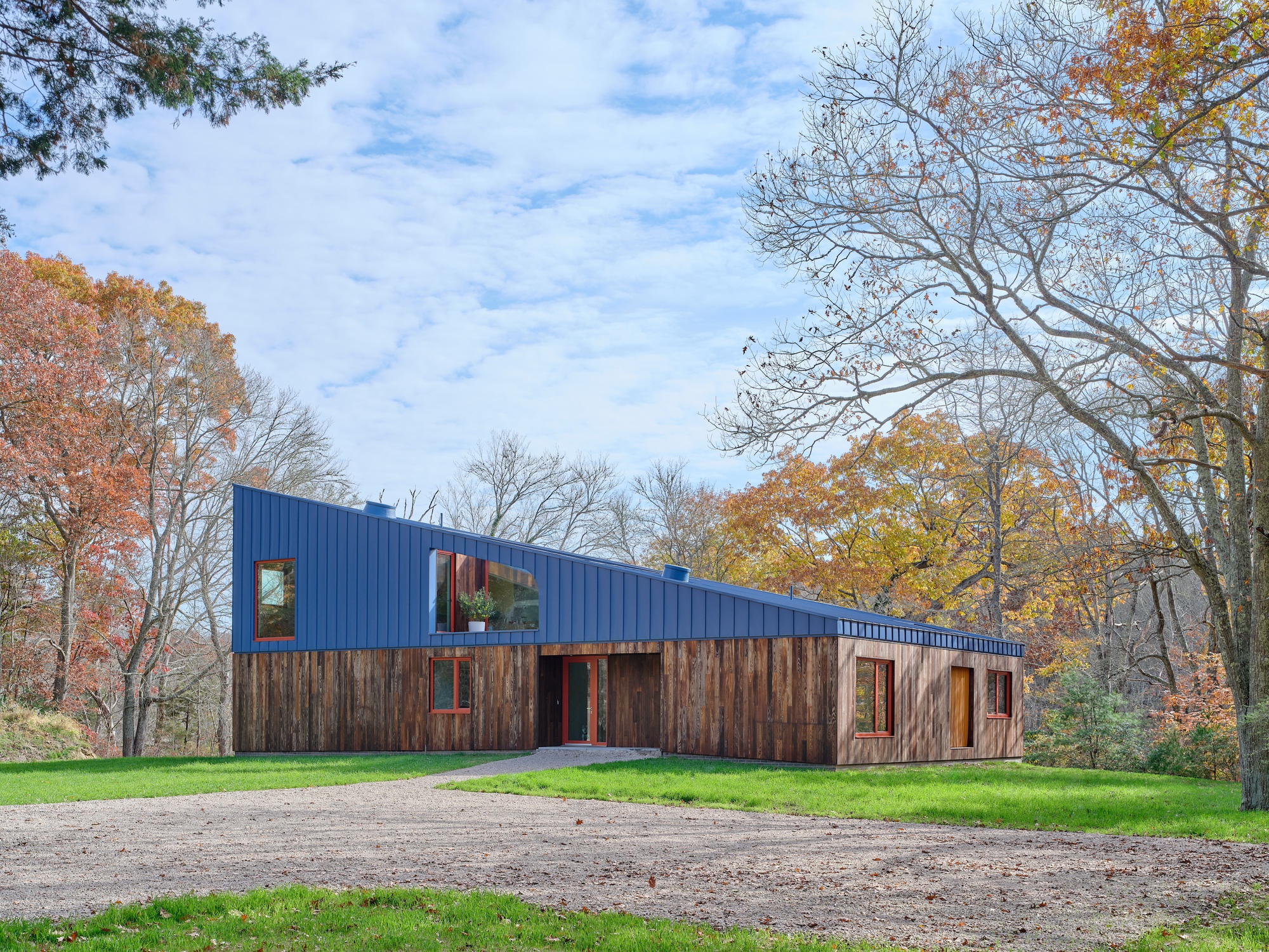 Two-volume residence with blue metal cladding above weathered timber siding set in a clearing among autumn trees