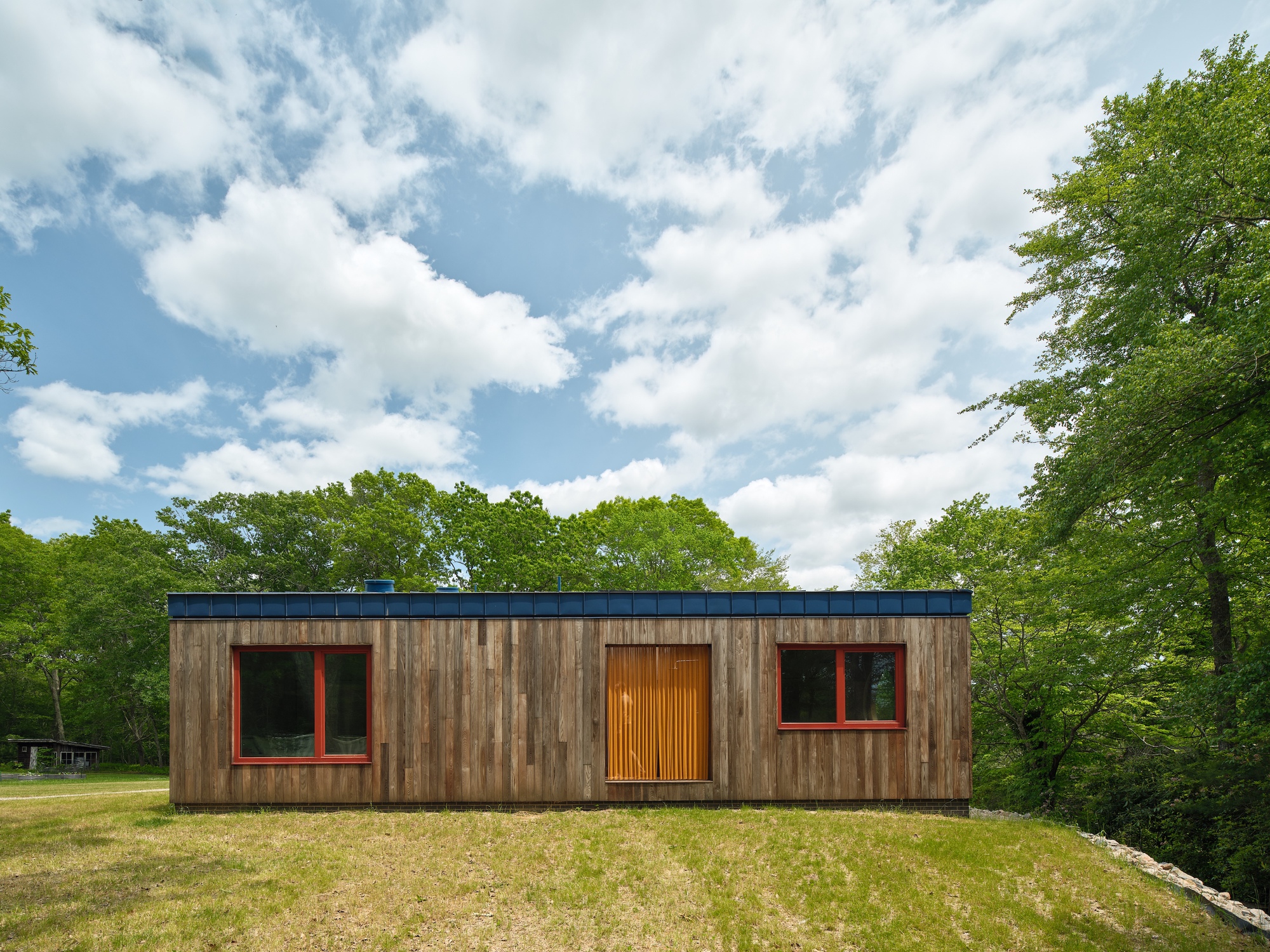 Garden facade showing weathered timber cladding with red-framed windows and vertical slat entry door under blue metal parapet