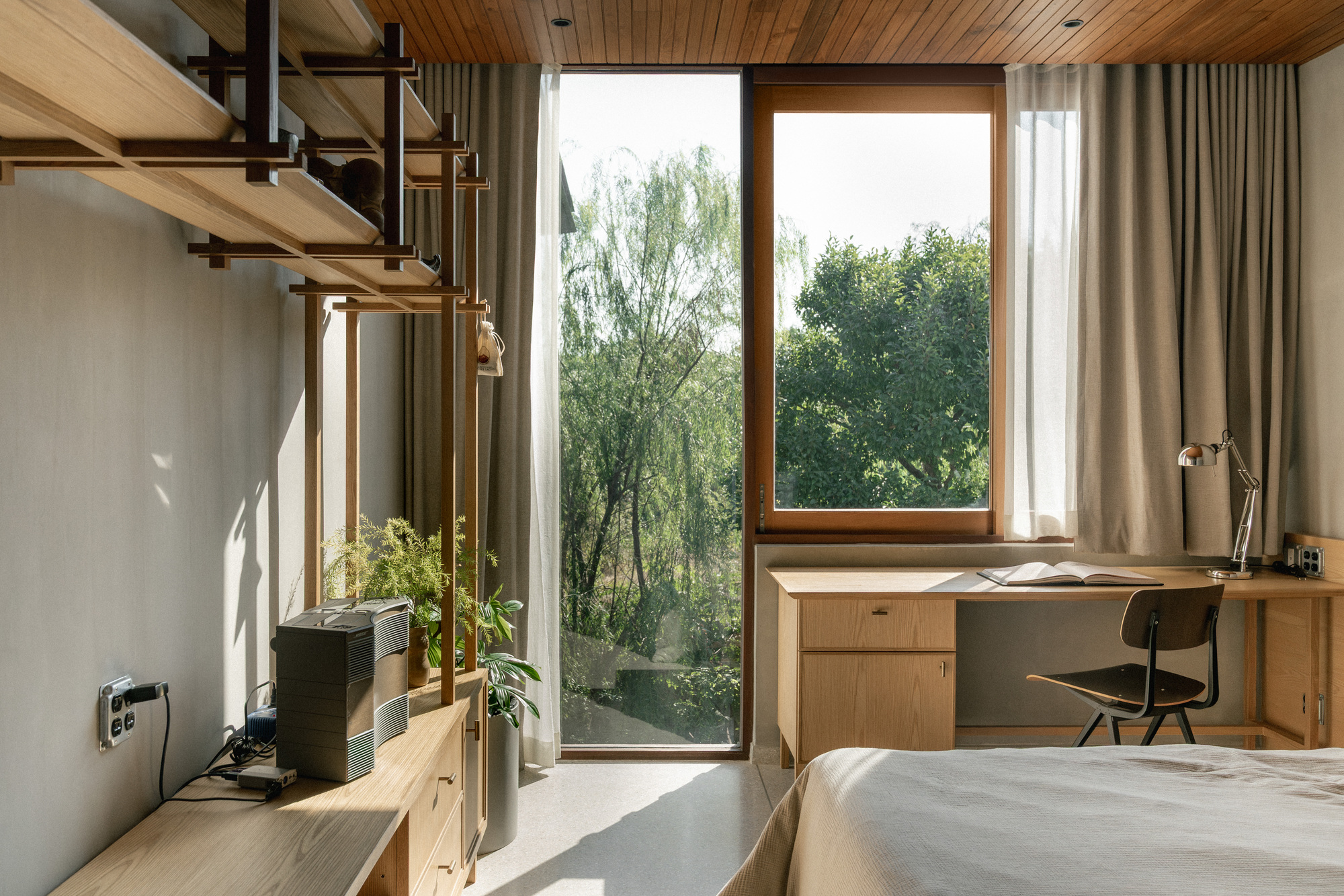 Bedroom with timber-lined ceiling, corner windows framing trees, and a built-in desk receiving morning light