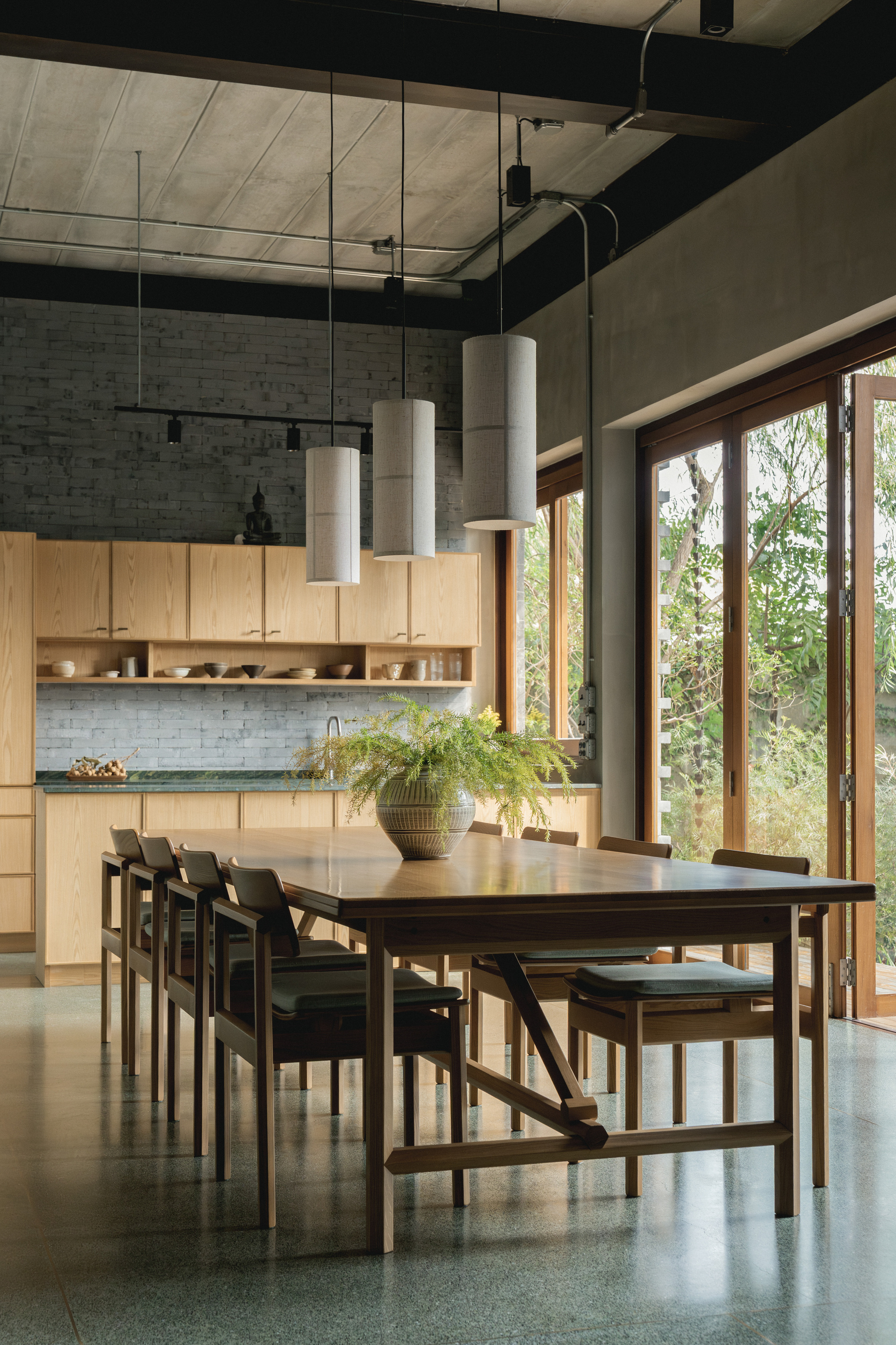 Dining area with exposed concrete ceiling, pendant lights above a timber table, and folding doors to greenery