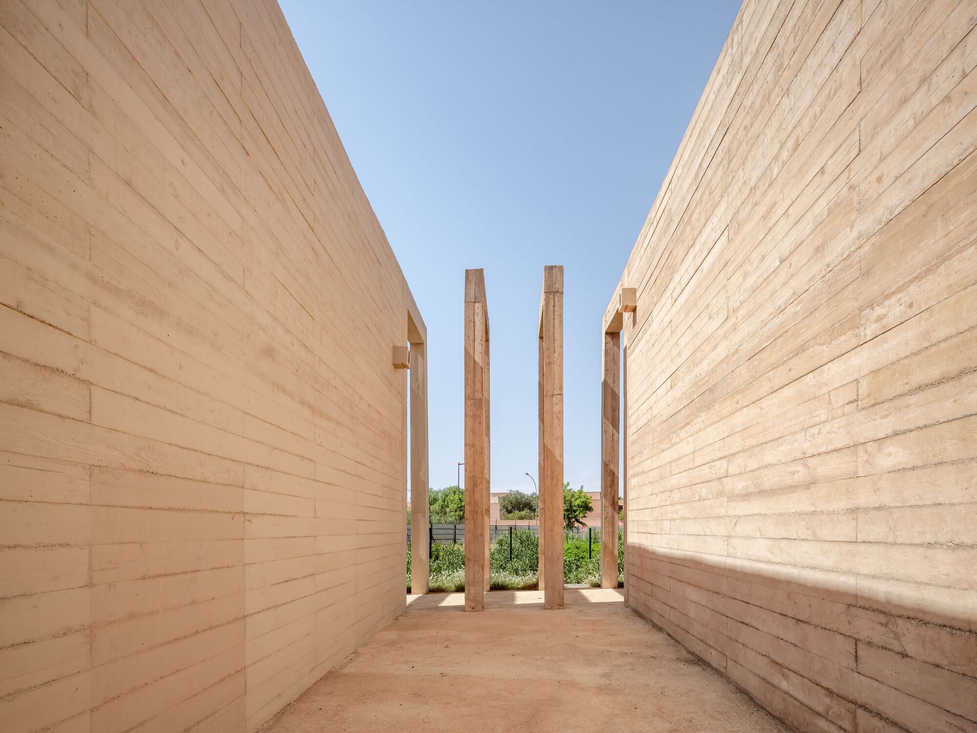 Narrow outdoor passage between rammed earth walls with four free-standing timber columns framing sky
