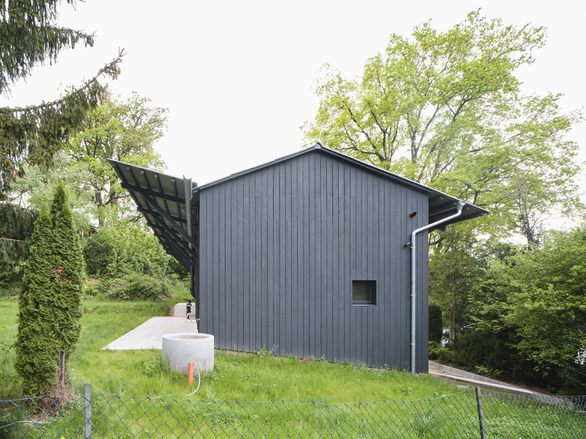 Gabled volume clad in vertical dark grey timber siding with extended eaves and adjacent white utility block