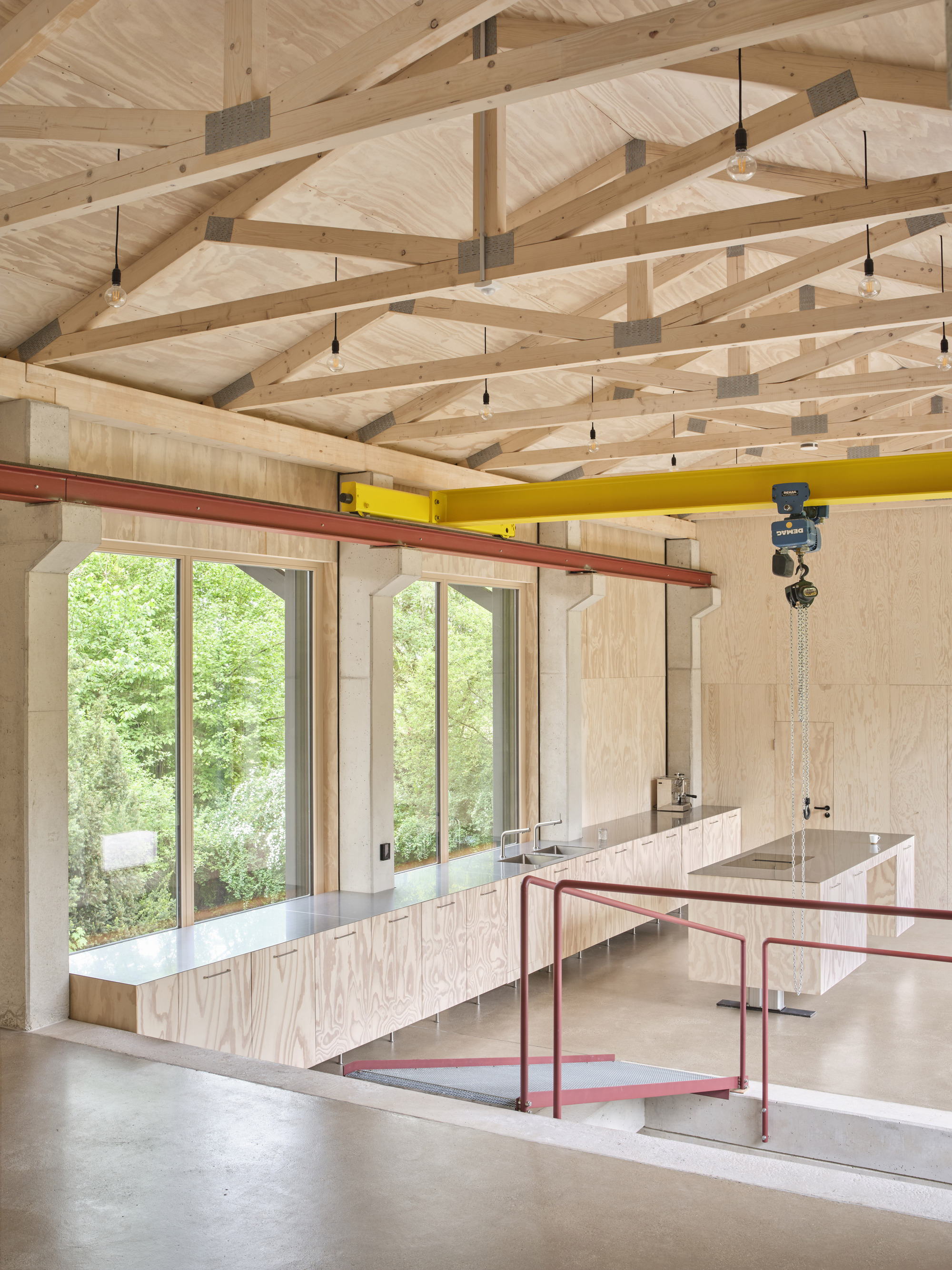 Interior view of exposed timber beam ceiling with red steel crane beam and plywood workspace overlooking trees
