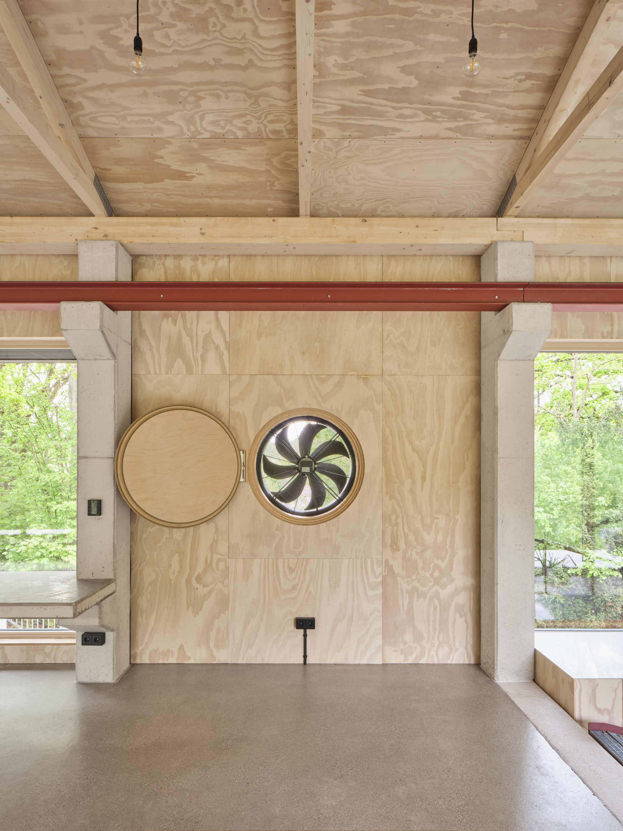 Plywood wall with two circular porthole windows and red steel beam beneath exposed timber ceiling