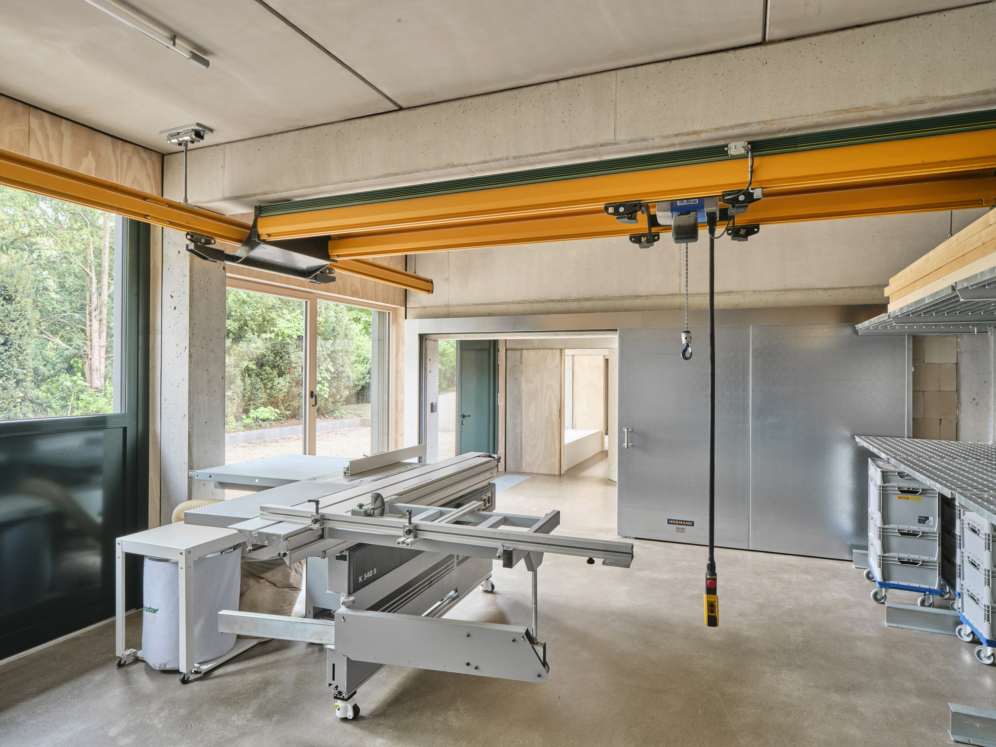 Workshop interior with yellow overhead crane beam and table saw facing plywood-framed glazing