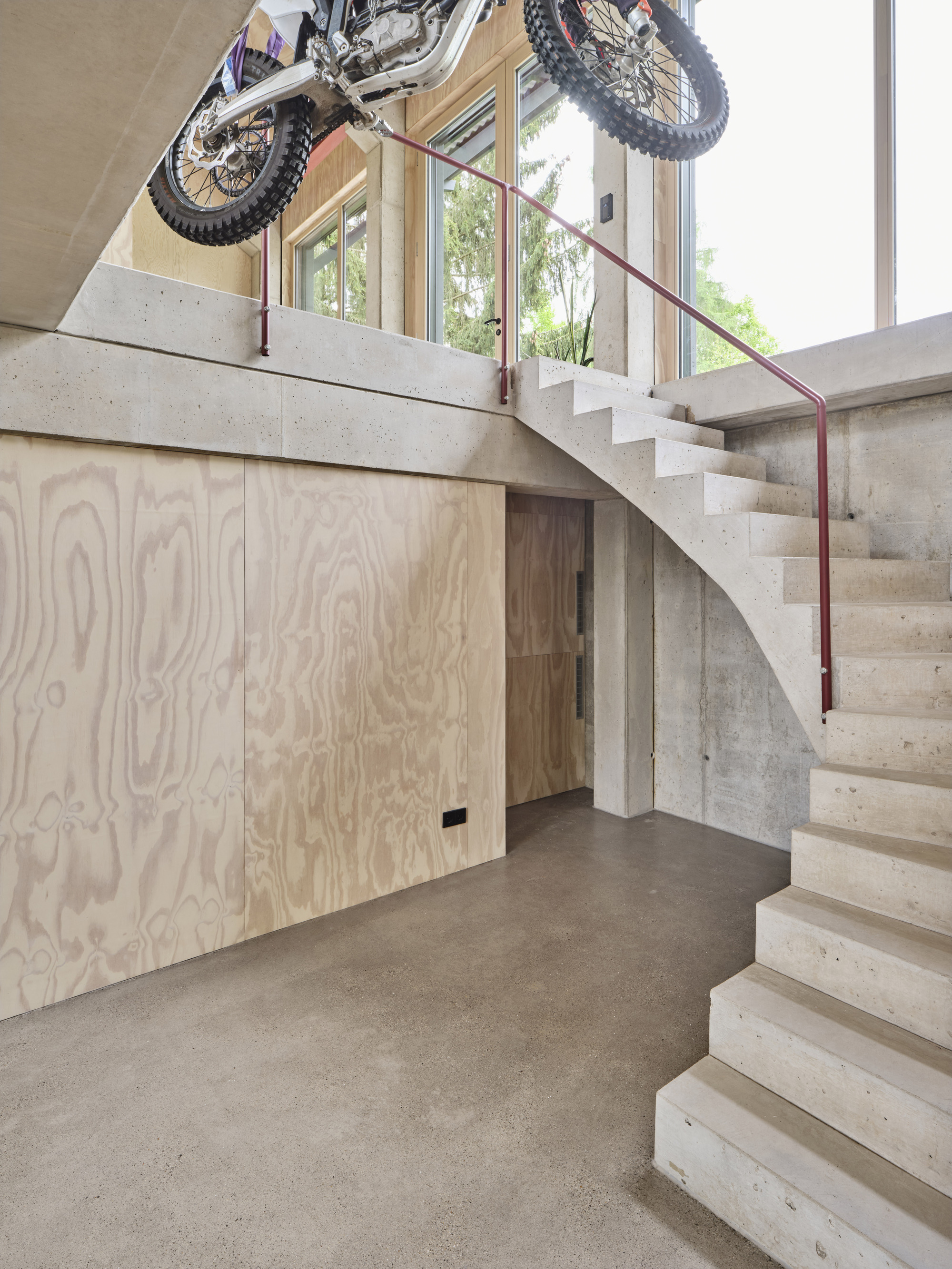 Concrete staircase with red steel handrail and plywood storage beneath displaying suspended motorcycle overhead
