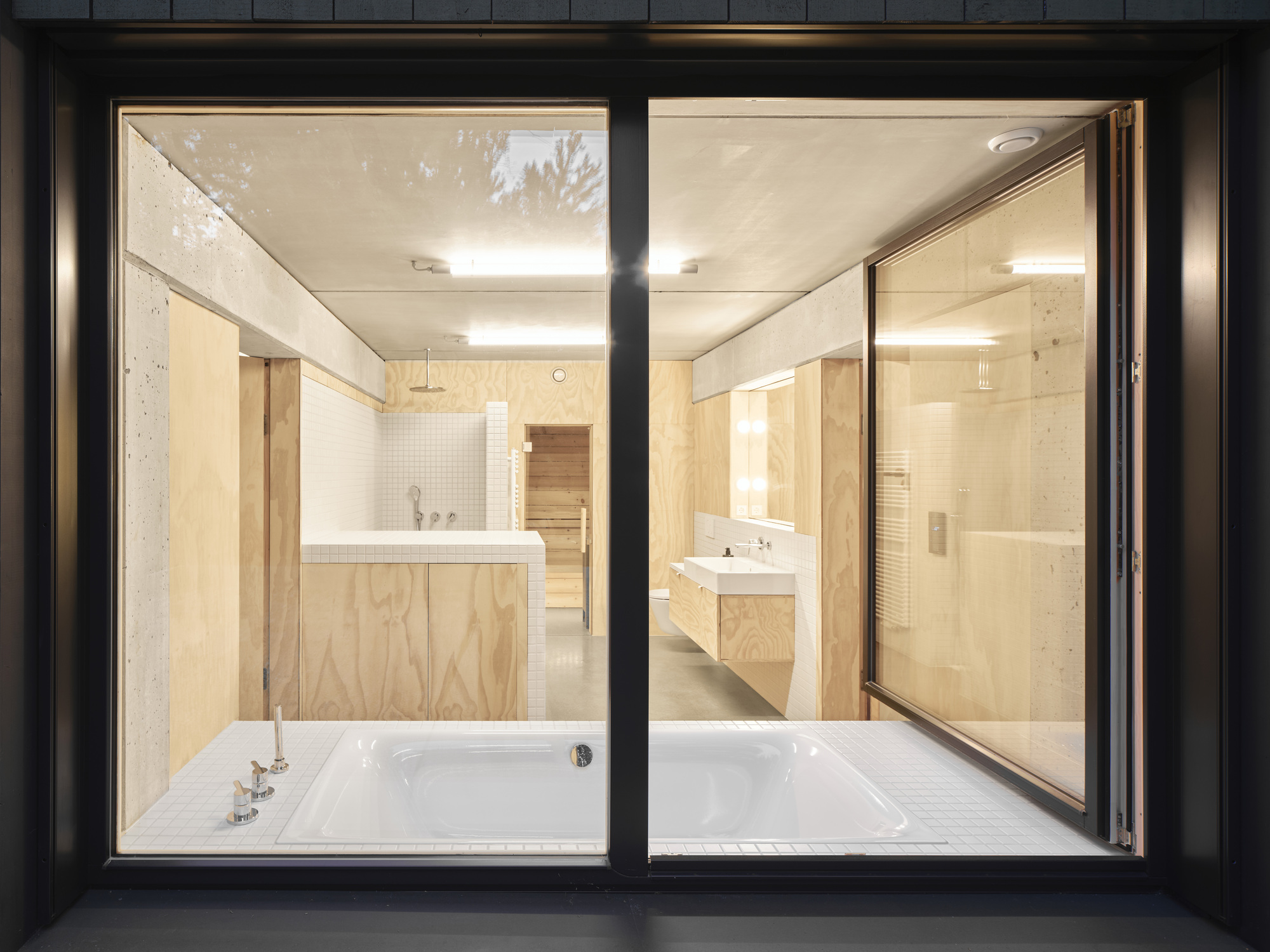 Night view through black metal window frames into bathroom with white tub and plywood cabinetry