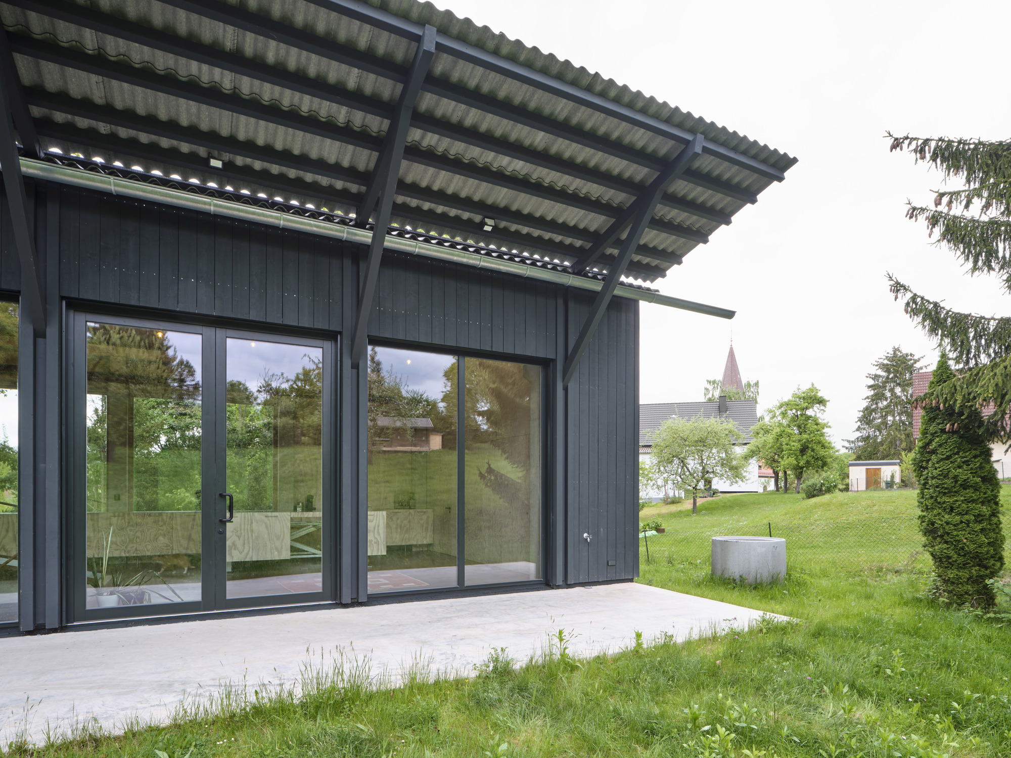 Dark timber-clad pavilion with large glass doors and corrugated metal canopy facing a grassy lawn