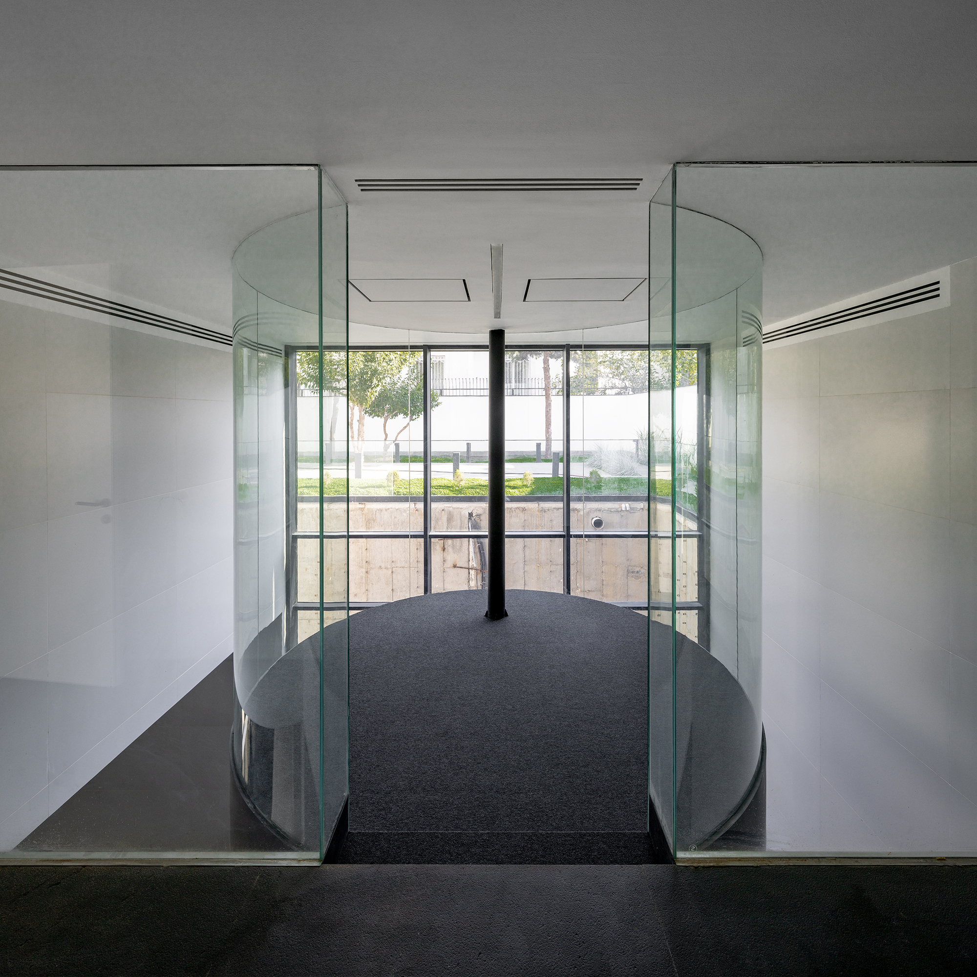 Glass-walled bridge with curved railings and central column overlooking a terraced garden courtyard