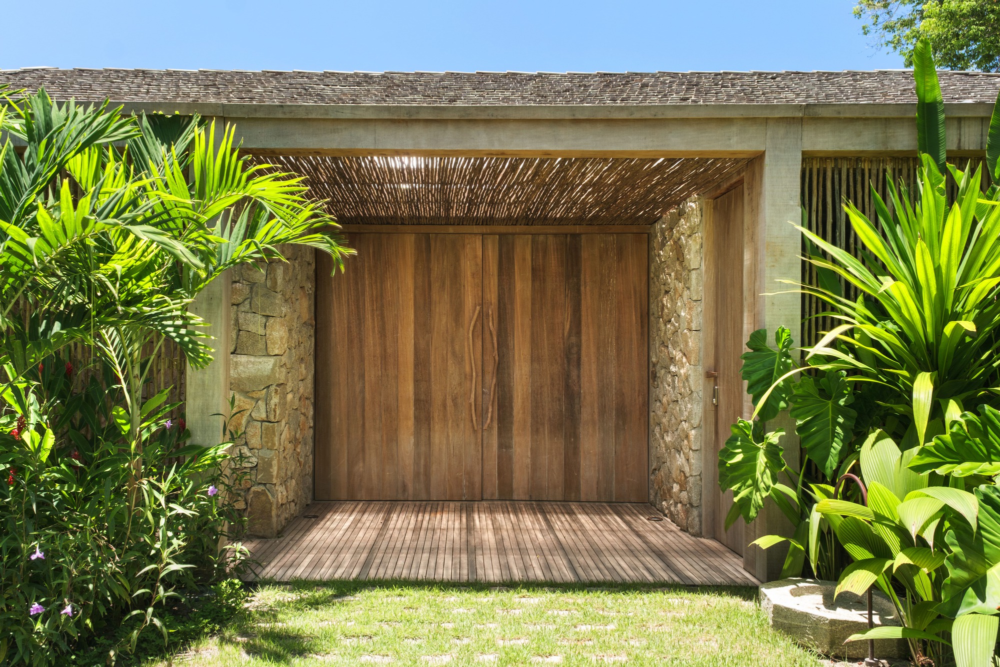 Covered entrance with timber doors, stone walls and woven wood ceiling surrounded by tropical plantings