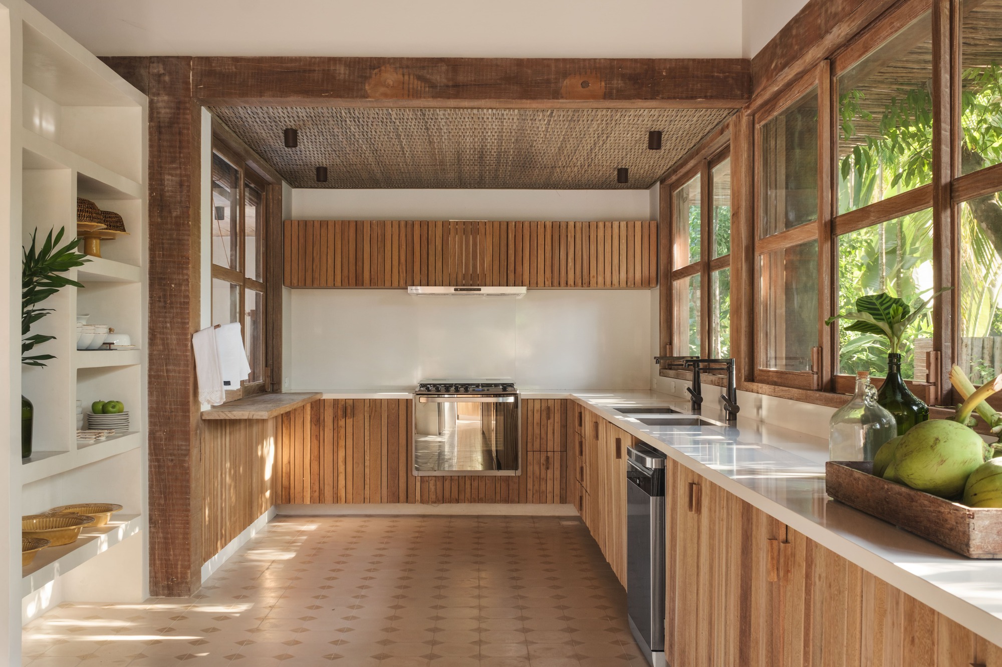 Kitchen with vertical timber cabinetry, woven ceiling panel and windows framing banana palms outside
