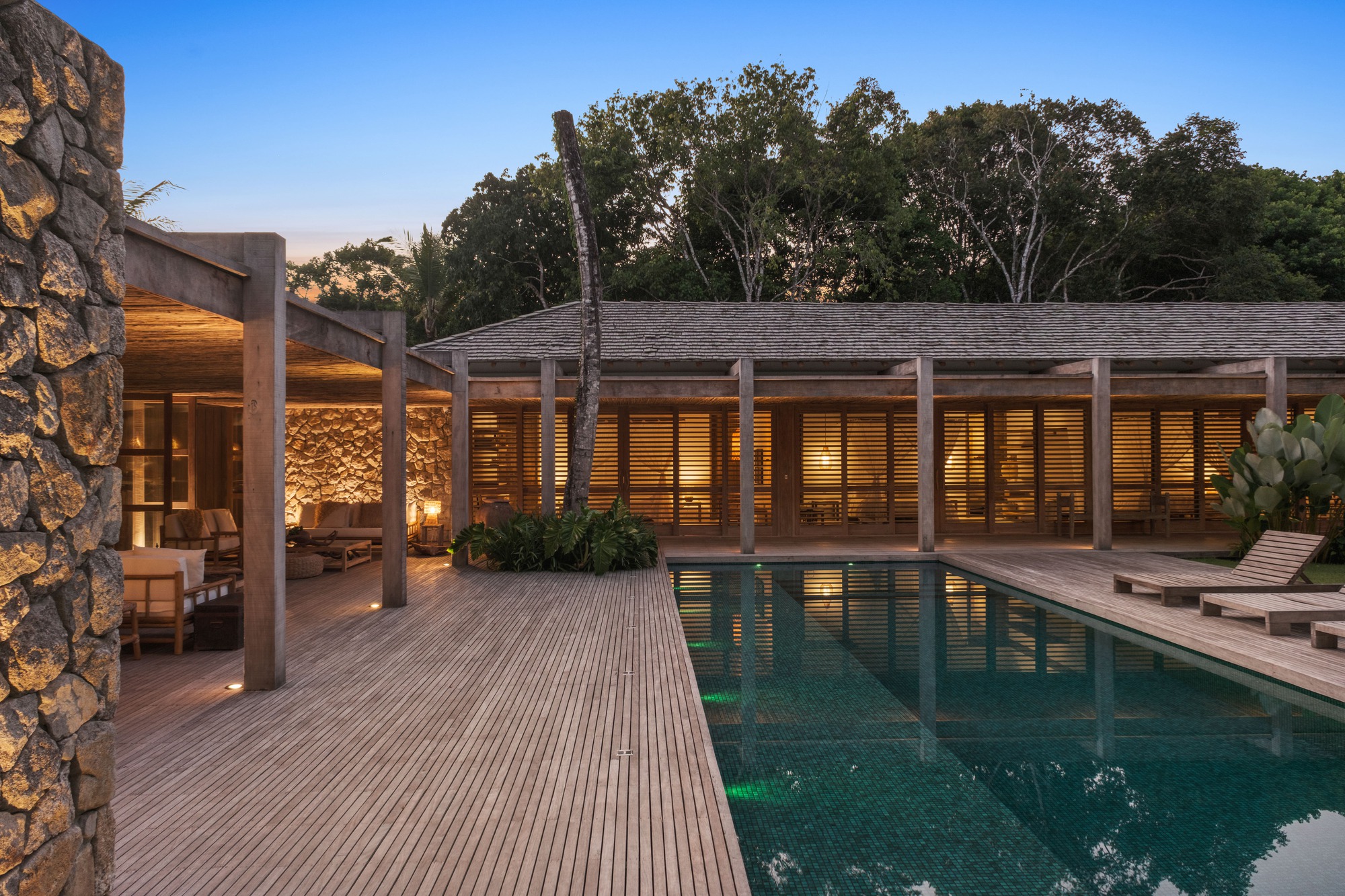 Pool terrace with timber decking and louvered facade wall illuminated at dusk under trees