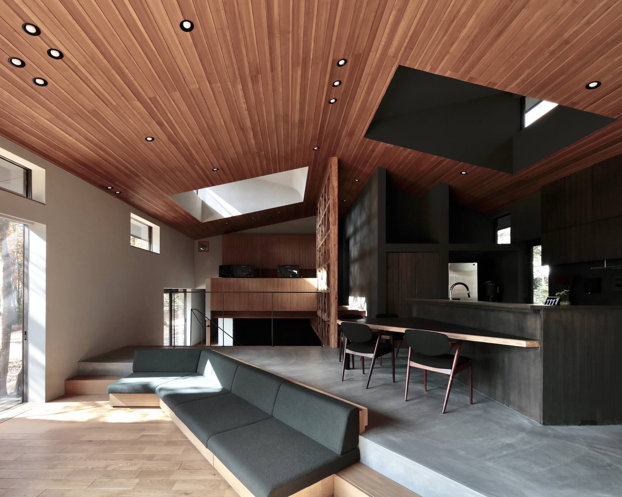 Double-height interior showing wood slat ceiling, built-in seating platform and dark timber kitchen volume