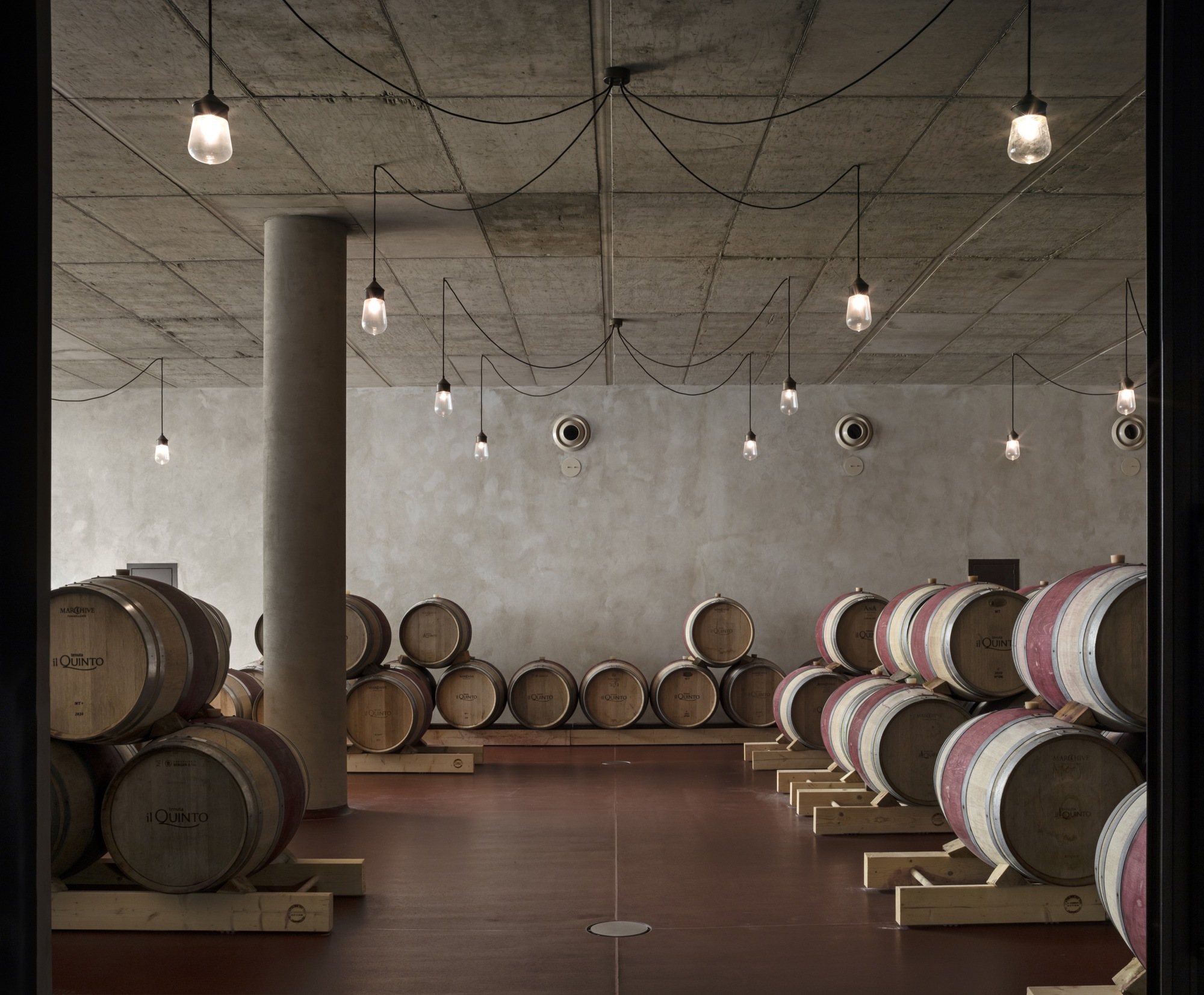 Barrel cellar with exposed concrete ceiling, pendant lights on draped cables, and wine casks on timber racks
