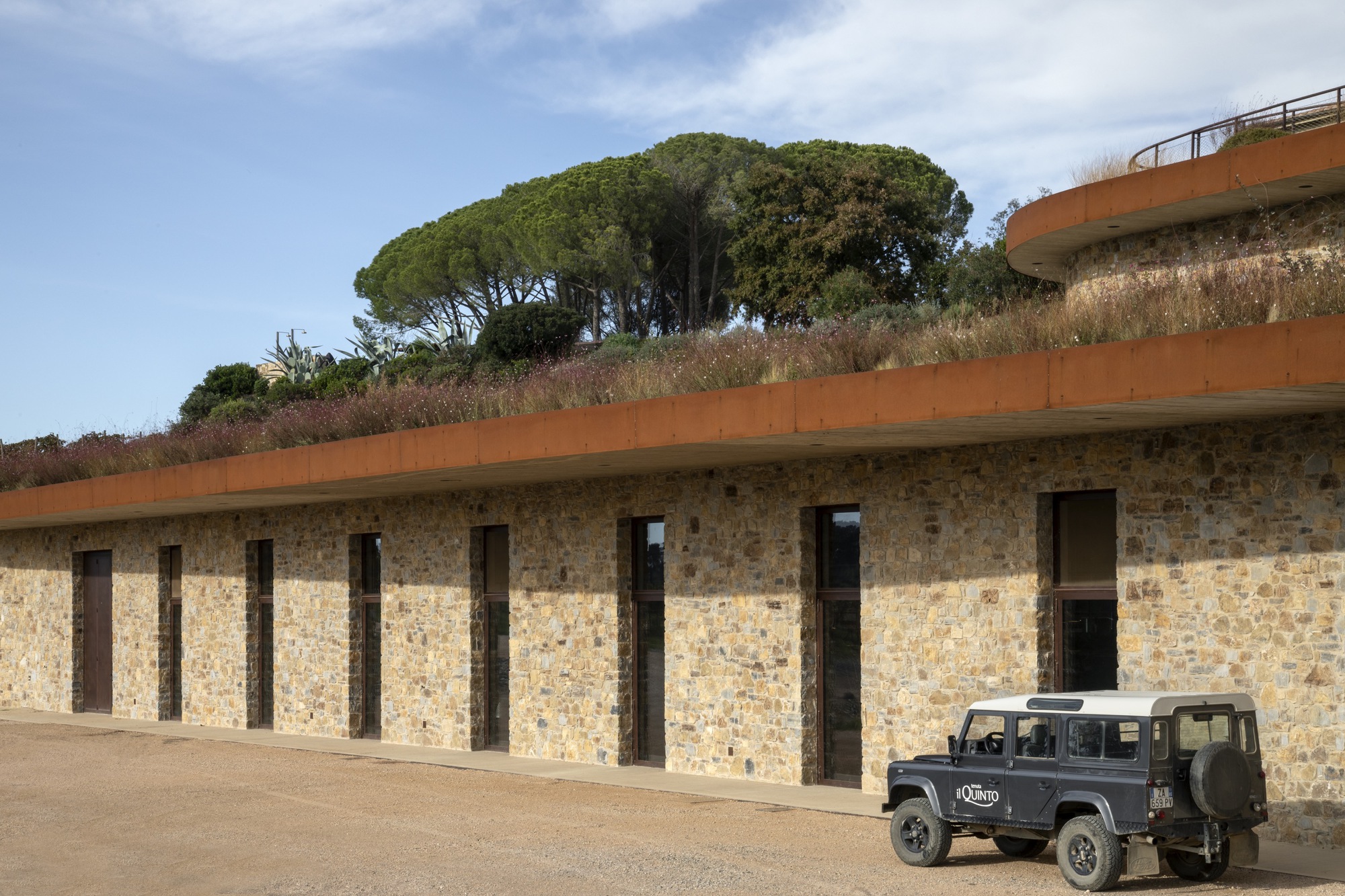 Stone facade with rhythmic timber-framed openings and planted roof edge with parked vehicle below