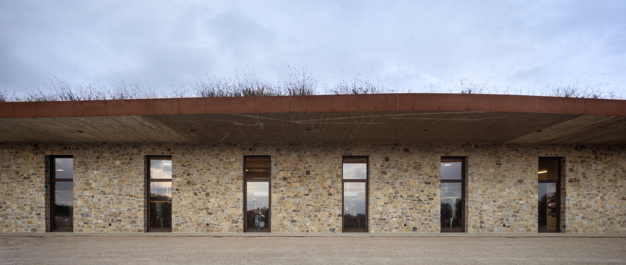 Stone facade with regularly spaced windows and timber door beneath overhanging planted roof
