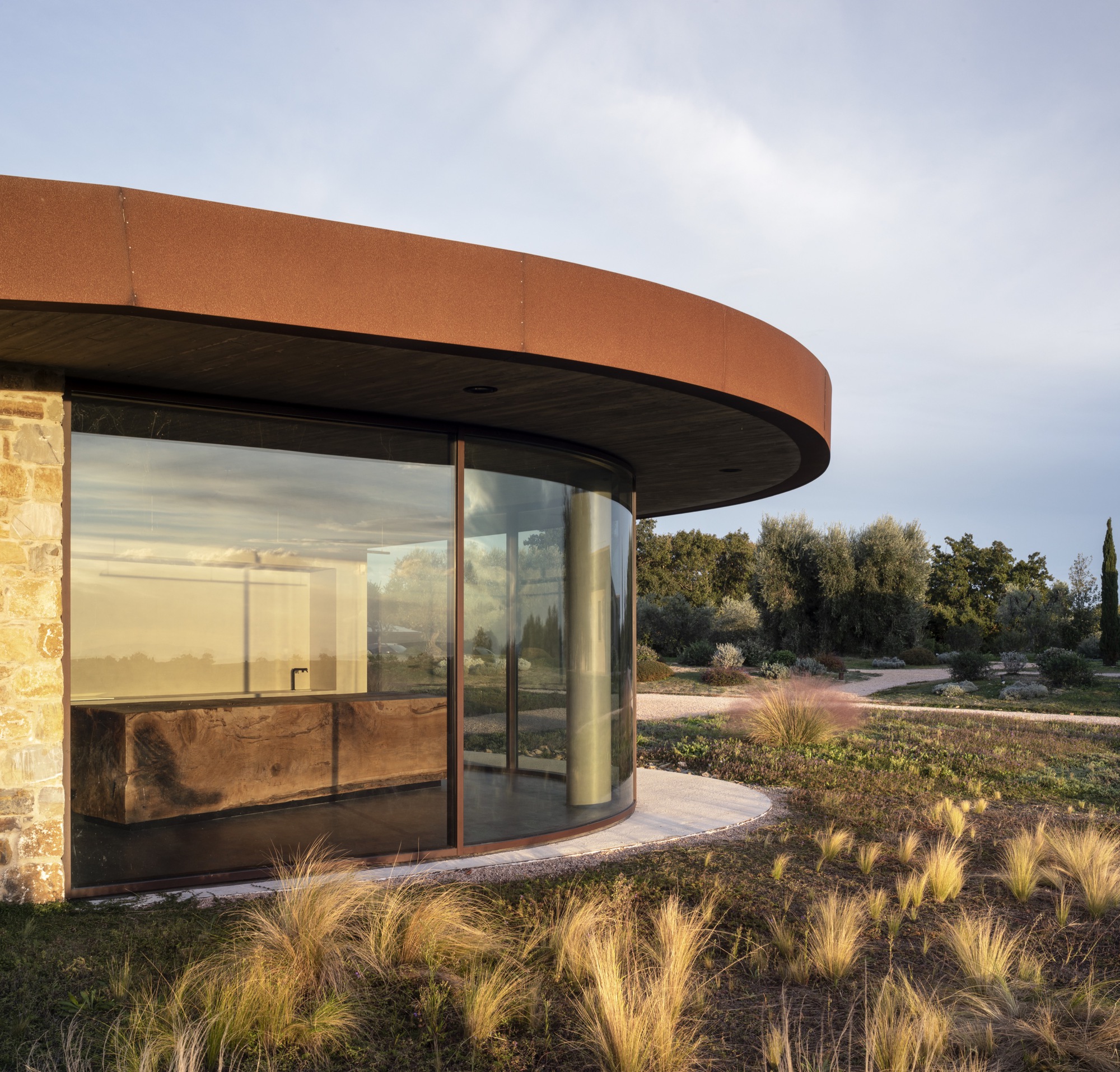 Curved glass facade with corten steel canopy and stone wall flanked by native grasses at dusk